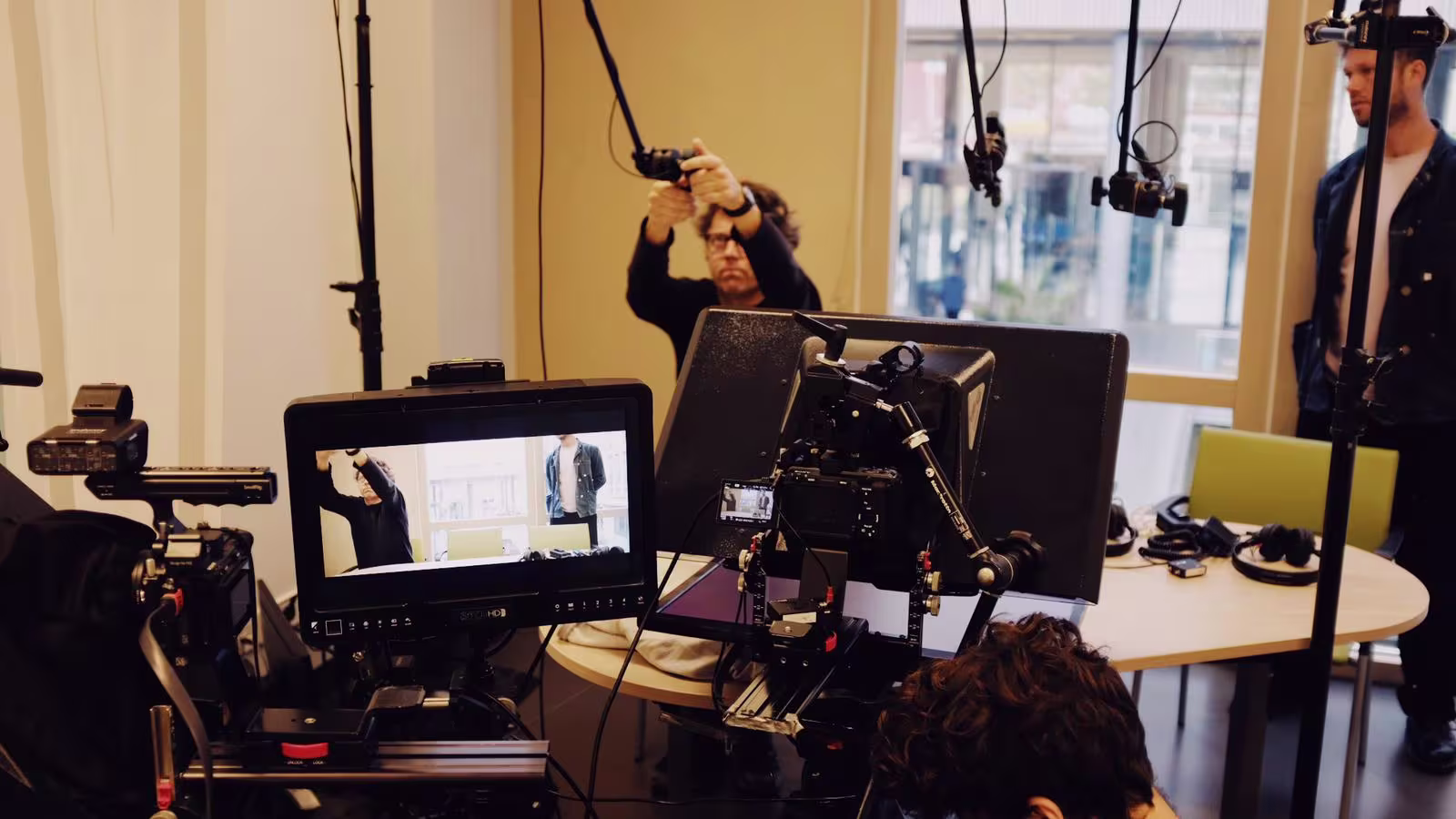 Een filmset met camera's, monitoren en crew die een opname voorbereiden in een lichte ruimte met grote ramen.