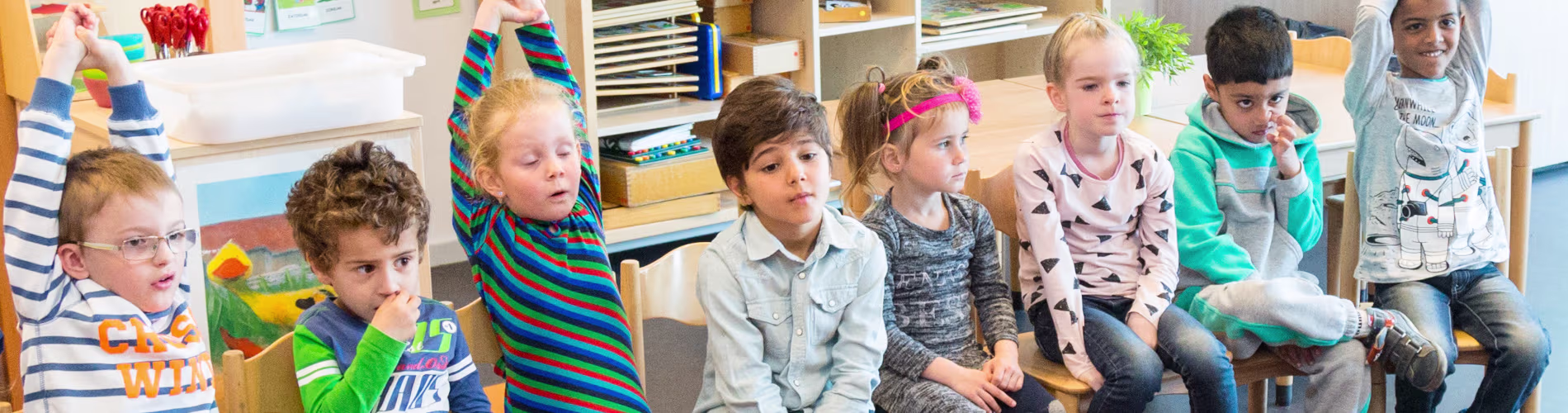Een groep kinderen zit in een klaslokaal, sommigen steken hun hand op tijdens een lesactiviteit.
