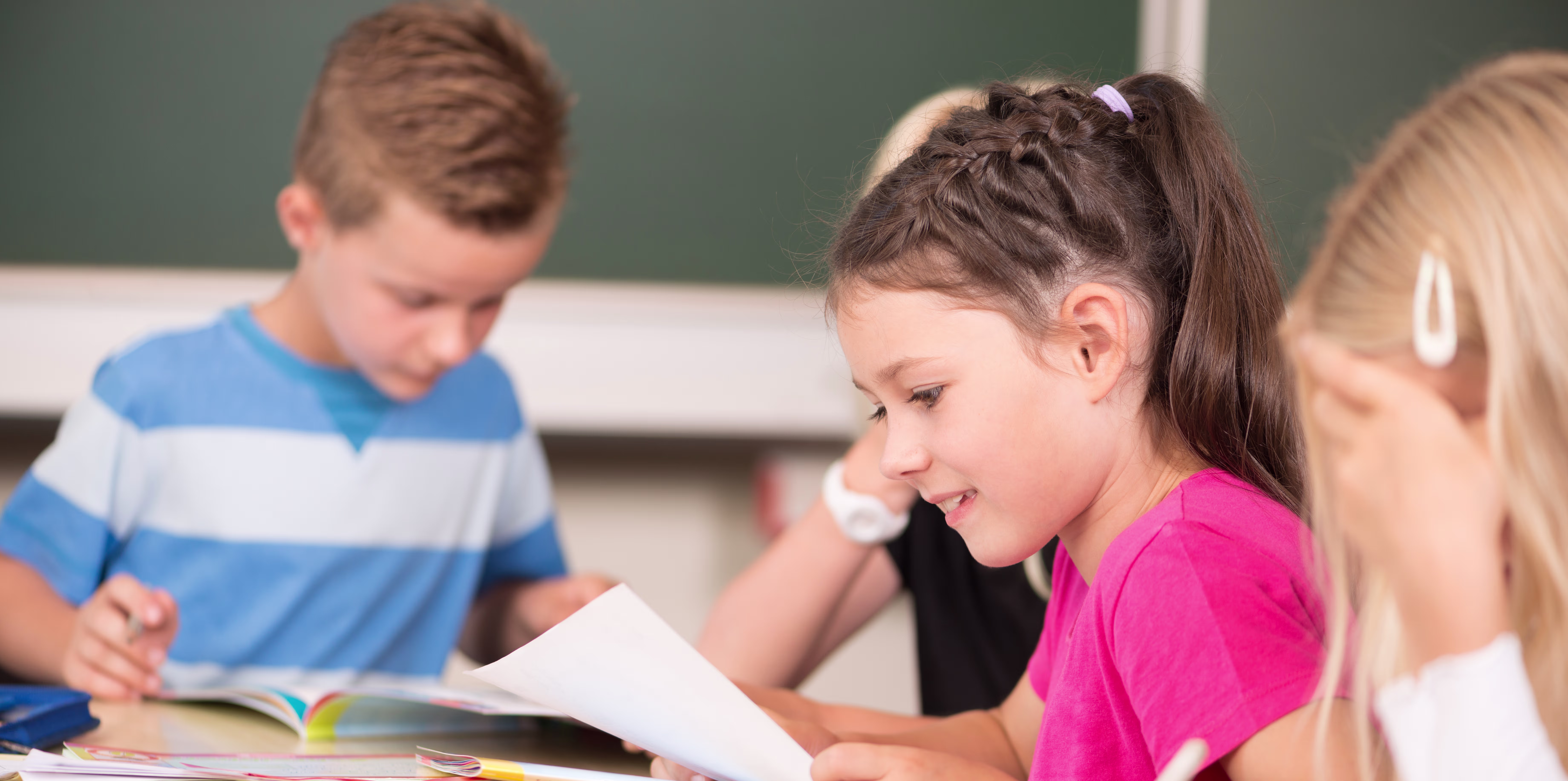 Drie leerlingen werken samen aan schoolwerk in een klaslokaal met groene schoolbord op de achtergrond.