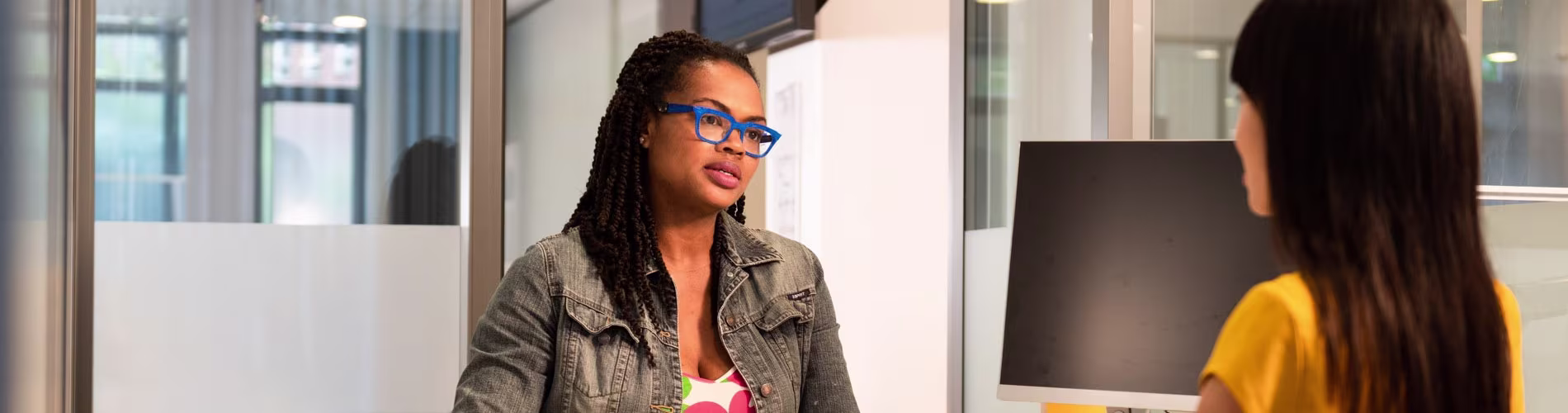 Twee vrouwen in gesprek op kantoor, een met blauwe bril en dreadlocks, de ander in gele trui bij computerscherm.
