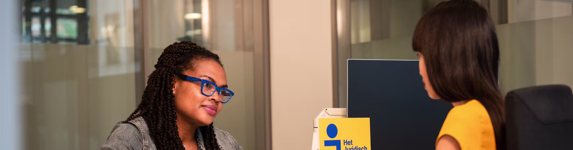 Twee vrouwen in gesprek op kantoor, een met bril en gevlochten haar, de ander in geel shirt bij Het Juridisch Loket logo.