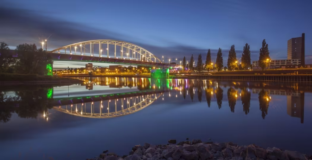 Verlichte John Frostbrug in Arnhem weerspiegelt in de rivier tijdens het blauwe uur, met stadsverlichting langs de kade.