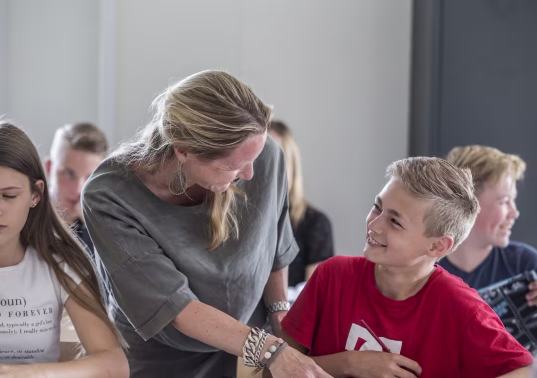 Een docente helpt een lachende leerling in een rood shirt tijdens de les, andere leerlingen zitten op de achtergrond.
