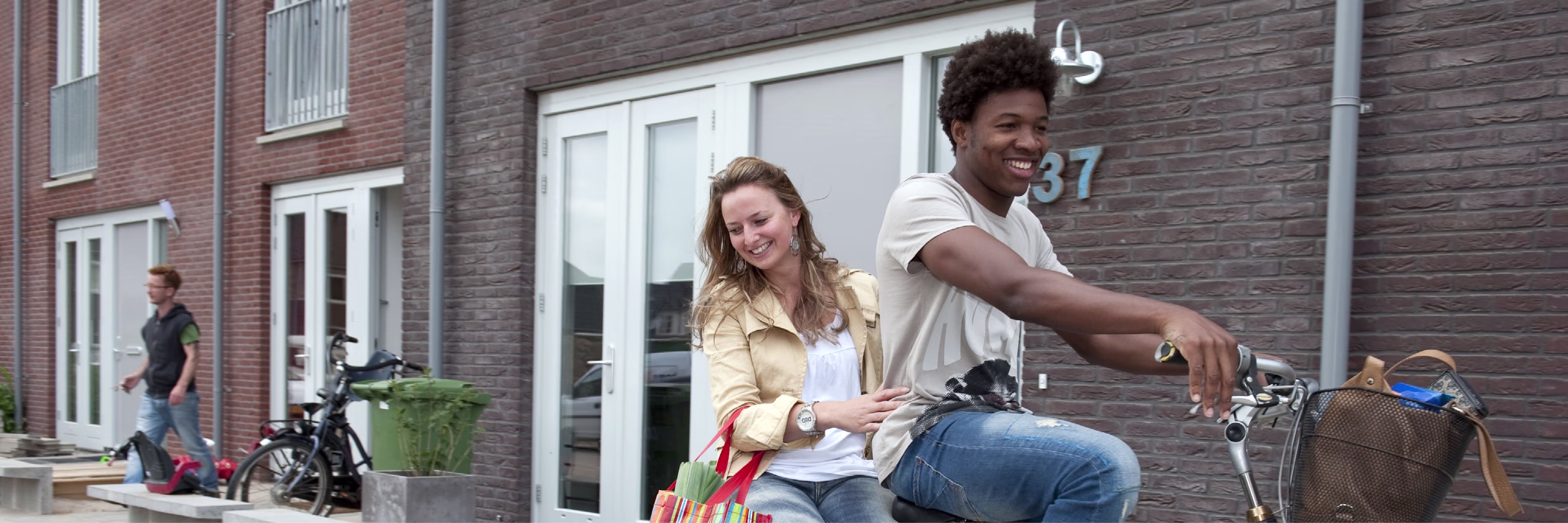 Twee lachende jongeren zitten op een fiets voor moderne woningen in een woonwijk.