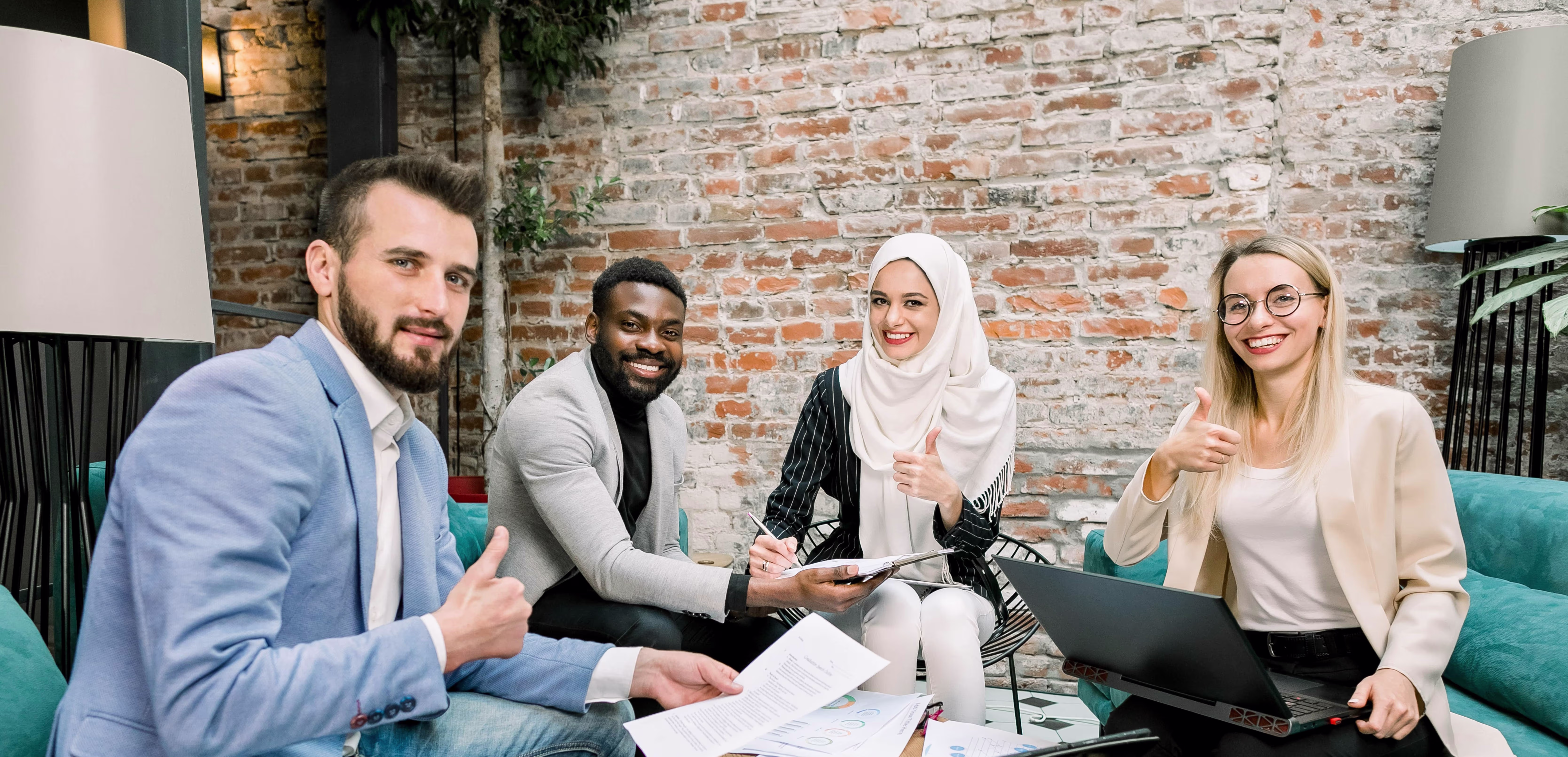 Vier diverse collega's zitten samen aan een tafel met laptop en documenten, allen met duim omhoog in moderne werkruimte.