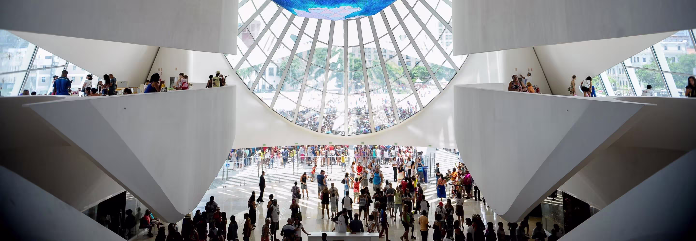 Bezoekers in modern museum met witte architectuur en groot glazen koepeldak waar natuurlijk licht doorheen valt.