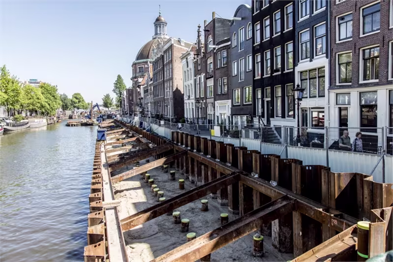 Bouwwerkzaamheden aan een Amsterdamse gracht met historische panden en kenmerkende architectuur op de achtergrond.