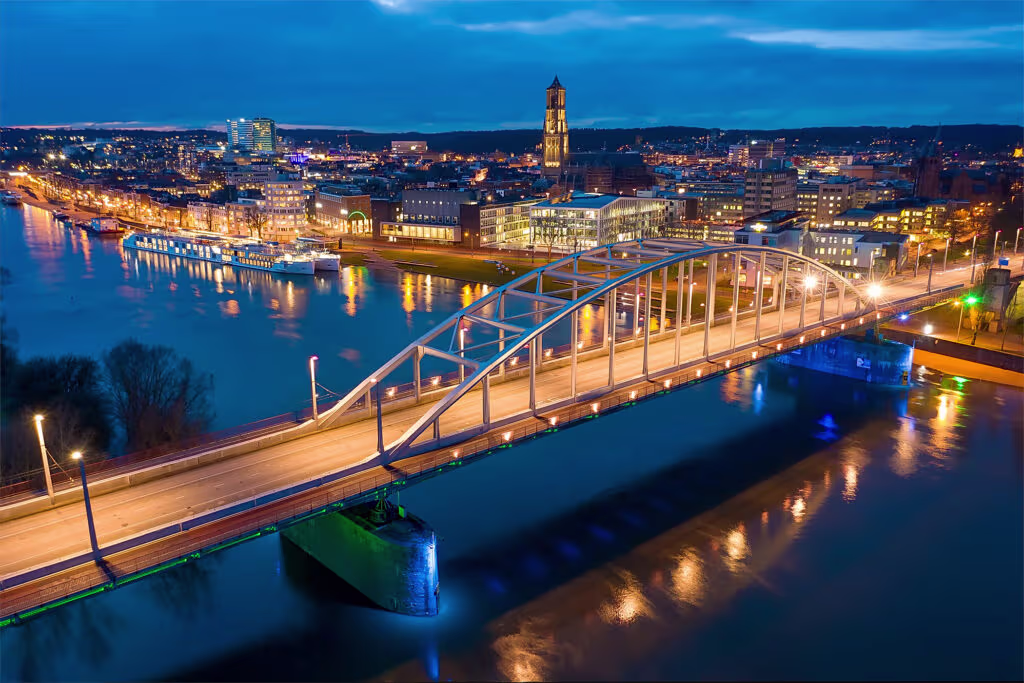 Een verlichte brug over de rivier met op de achtergrond de skyline van Arnhem bij avond met kleurrijke stadslichten.