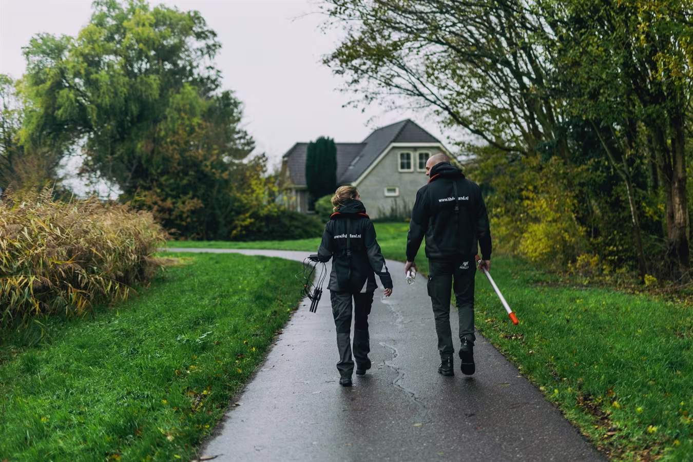 Twee zorgmedewerkers lopen over een landelijk pad naar een huis, één met een wandelstok.