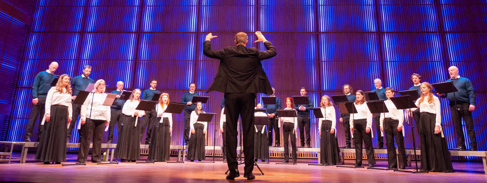 Een dirigent leidt een koor van zangers in zwart-witte kleding op een podium met blauwe verlichting.