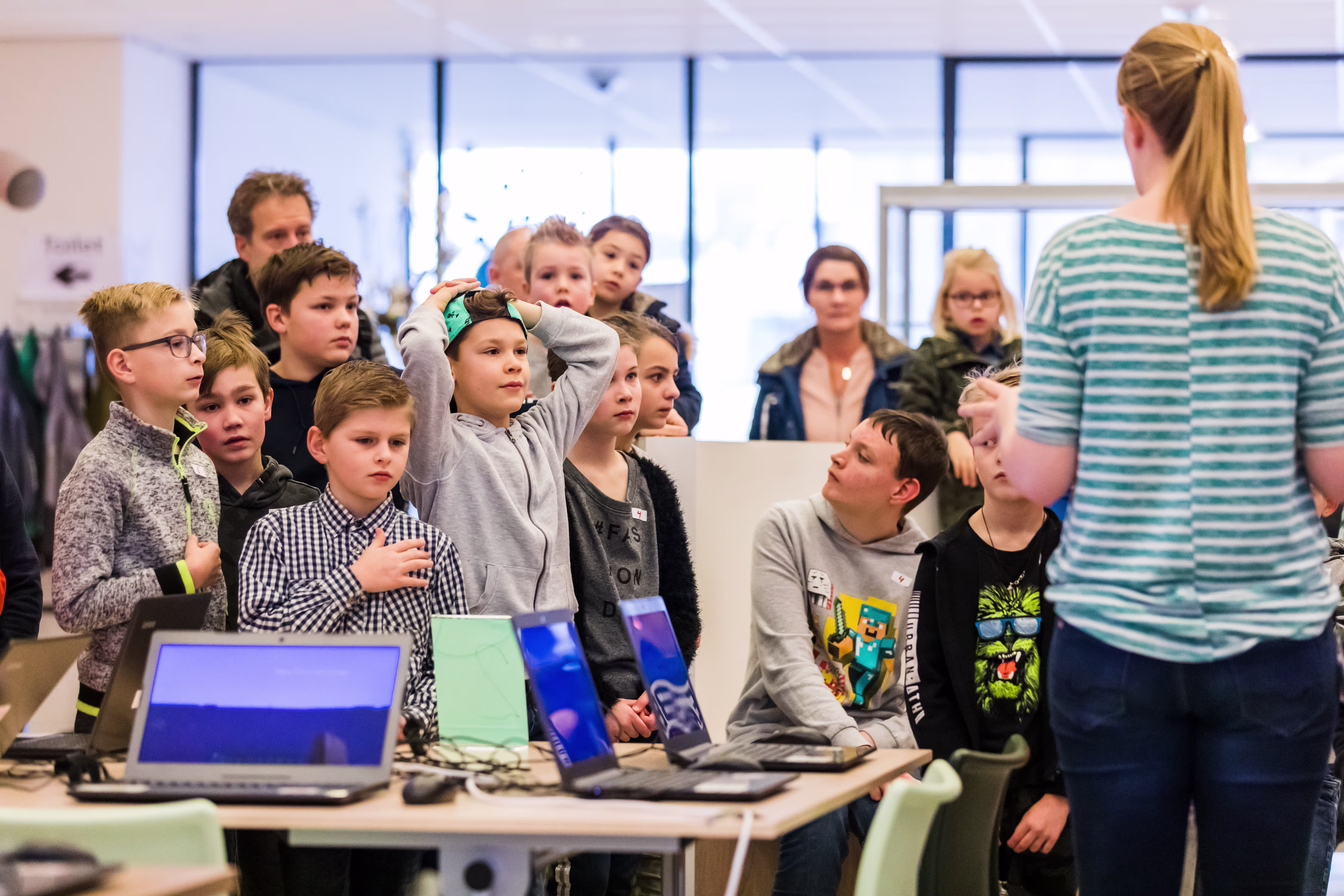 Een groep kinderen en jongeren kijkt aandachtig naar een vrouw die uitleg geeft bij computers in de bibliotheek.