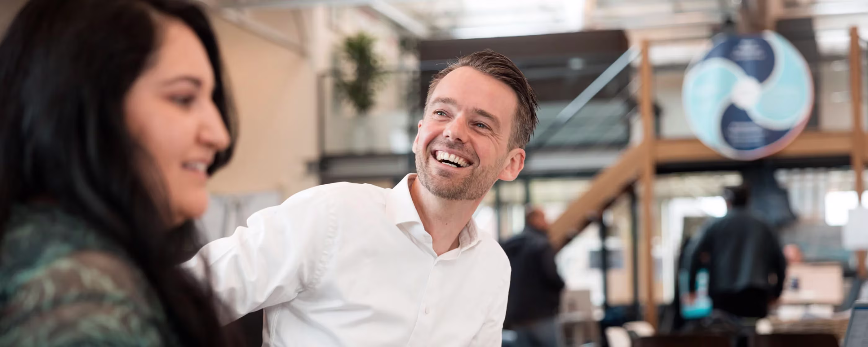 Twee lachende collega's in gesprek op een moderne kantoorwerkplek met houten kasten en planten op de achtergrond.