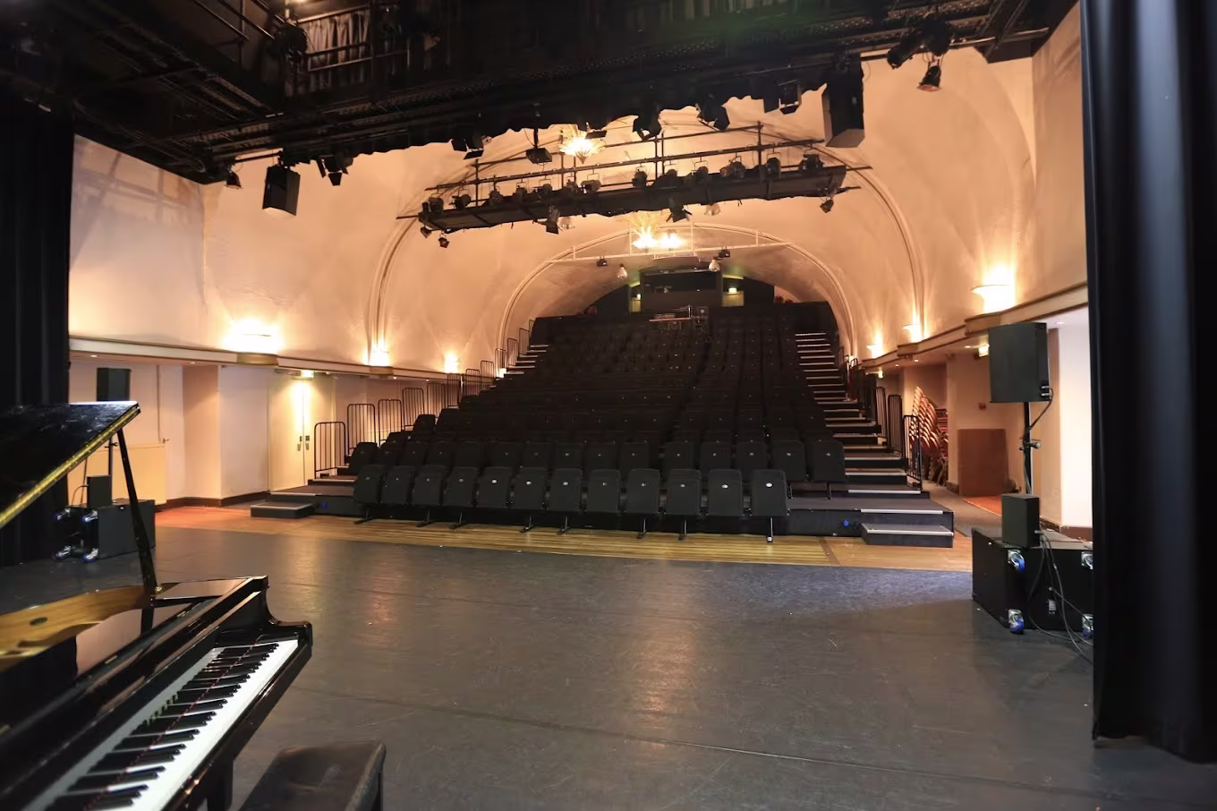 Interieur van een theater met podium, piano en tribunes met zitplaatsen onder warm verlichte gewelfde plafonds.