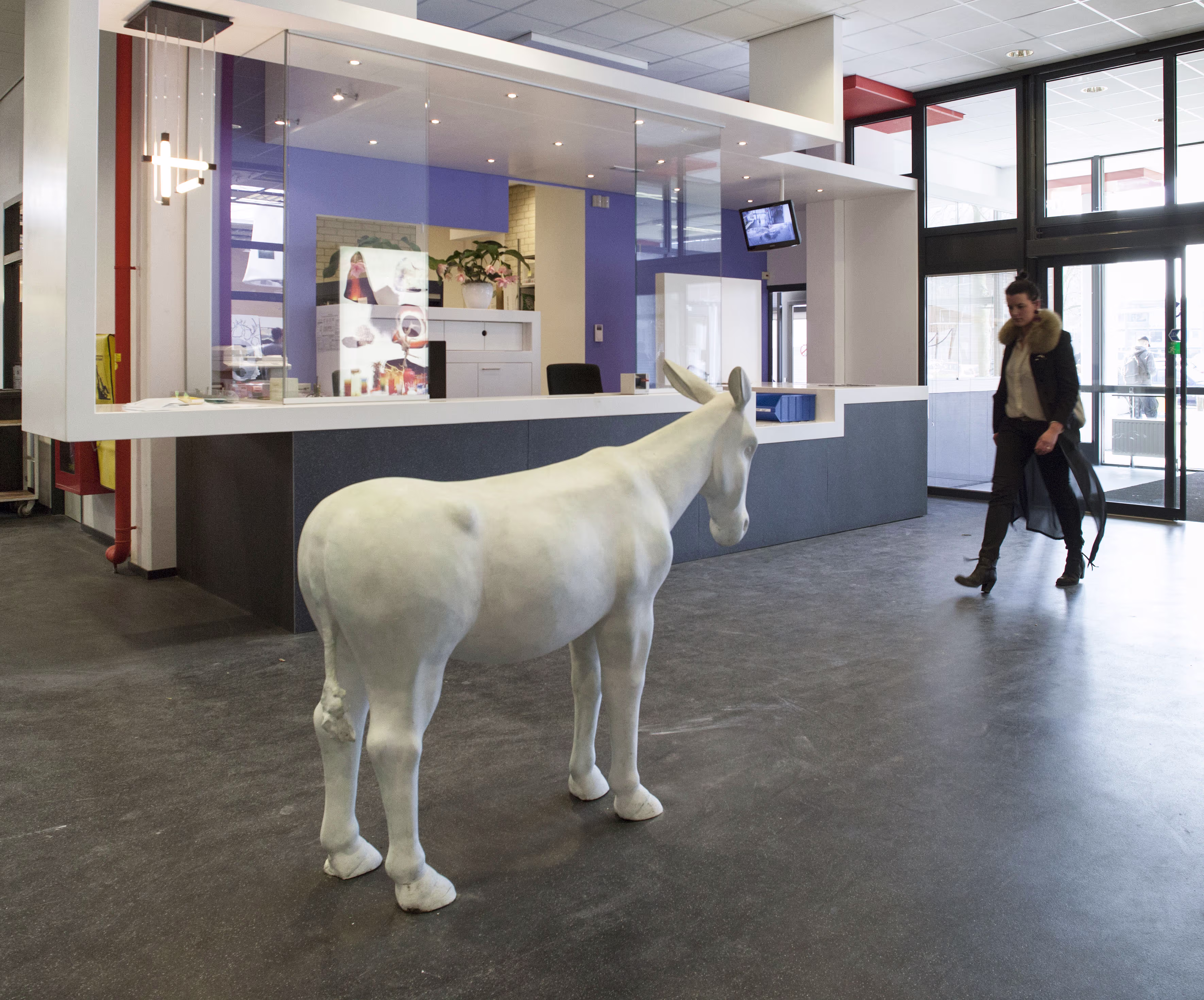 Een wit paardenbeeld staat in een moderne receptieruimte terwijl een persoon op de achtergrond loopt.