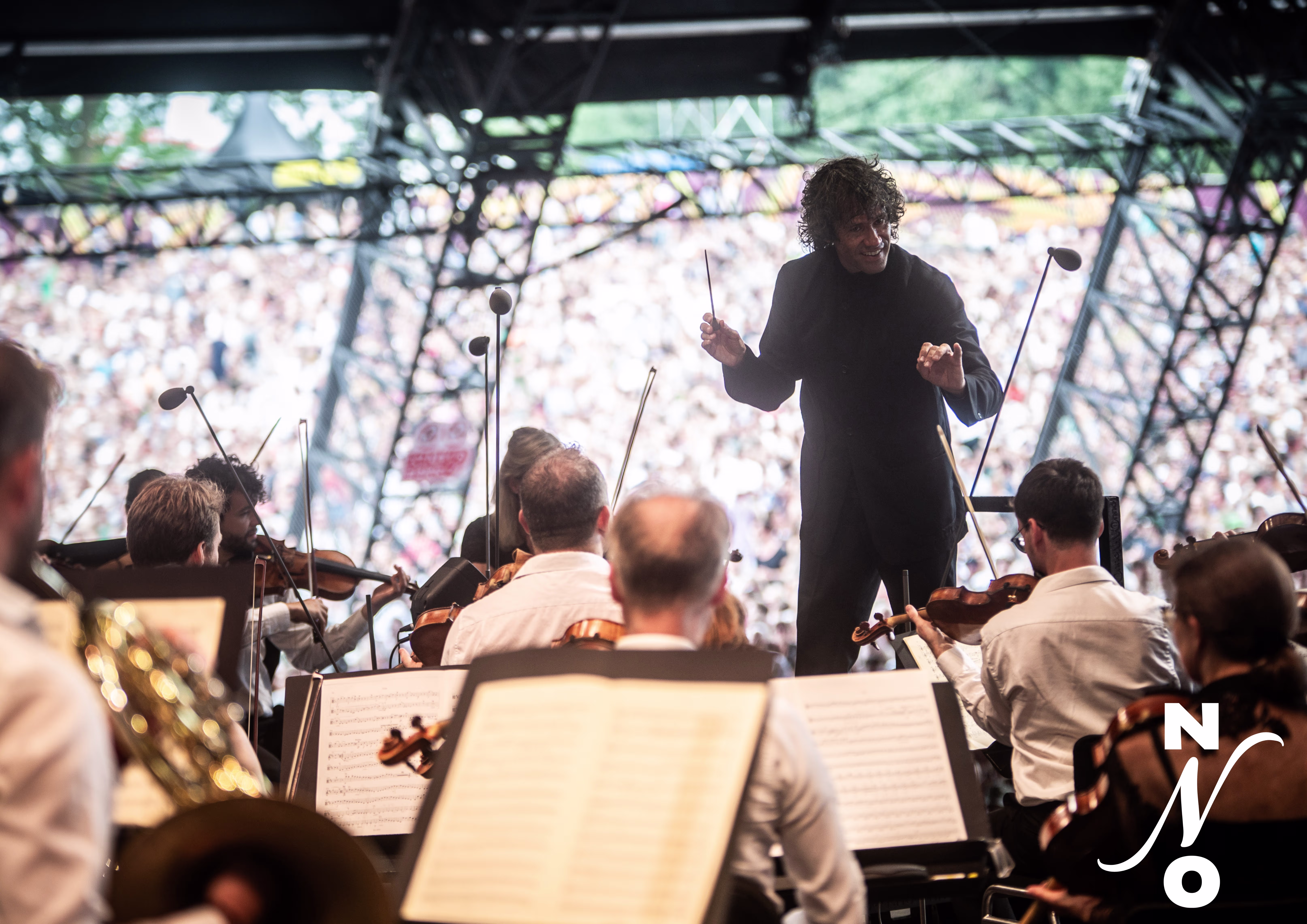 Een dirigent leidt het orkest tijdens een uitvoering in een grote concertzaal met publiek op de achtergrond.
