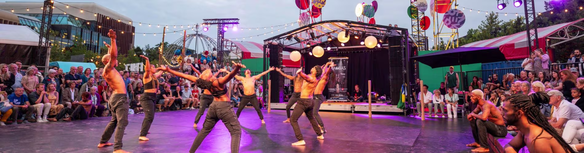 Dansers in oranje shirts treden op voor een grote menigte tijdens een openlucht theaterfestival met kleurrijke verlichting.
