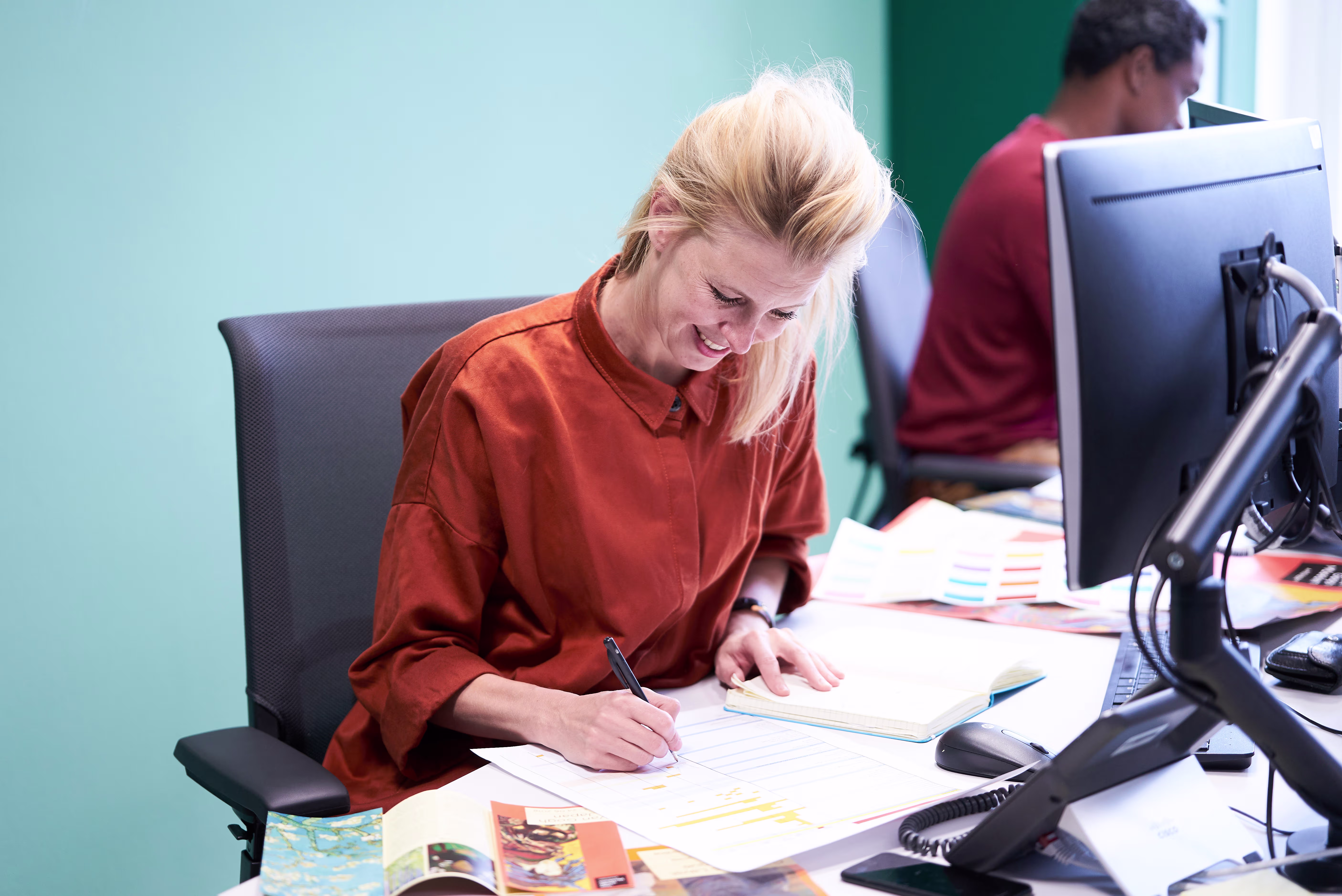 Een blonde vrouw in oranje blouse schrijft lachend aan een bureau met computer en telefoon in een modern kantoor.
