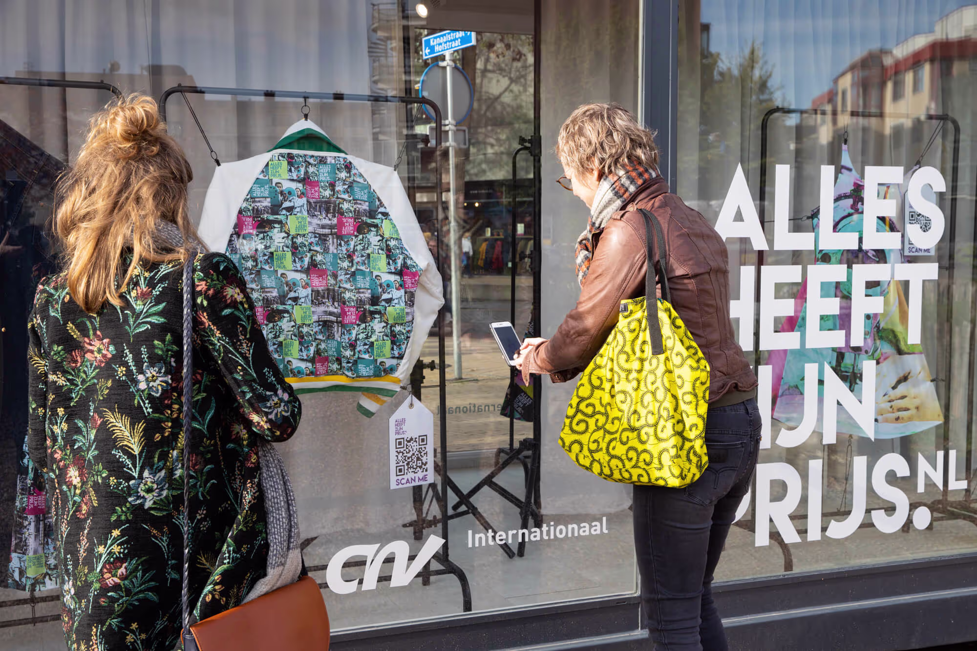 Twee vrouwen bekijken een kleurrijke trui in de etalage van een winkel met CNV Internationaal logo.
