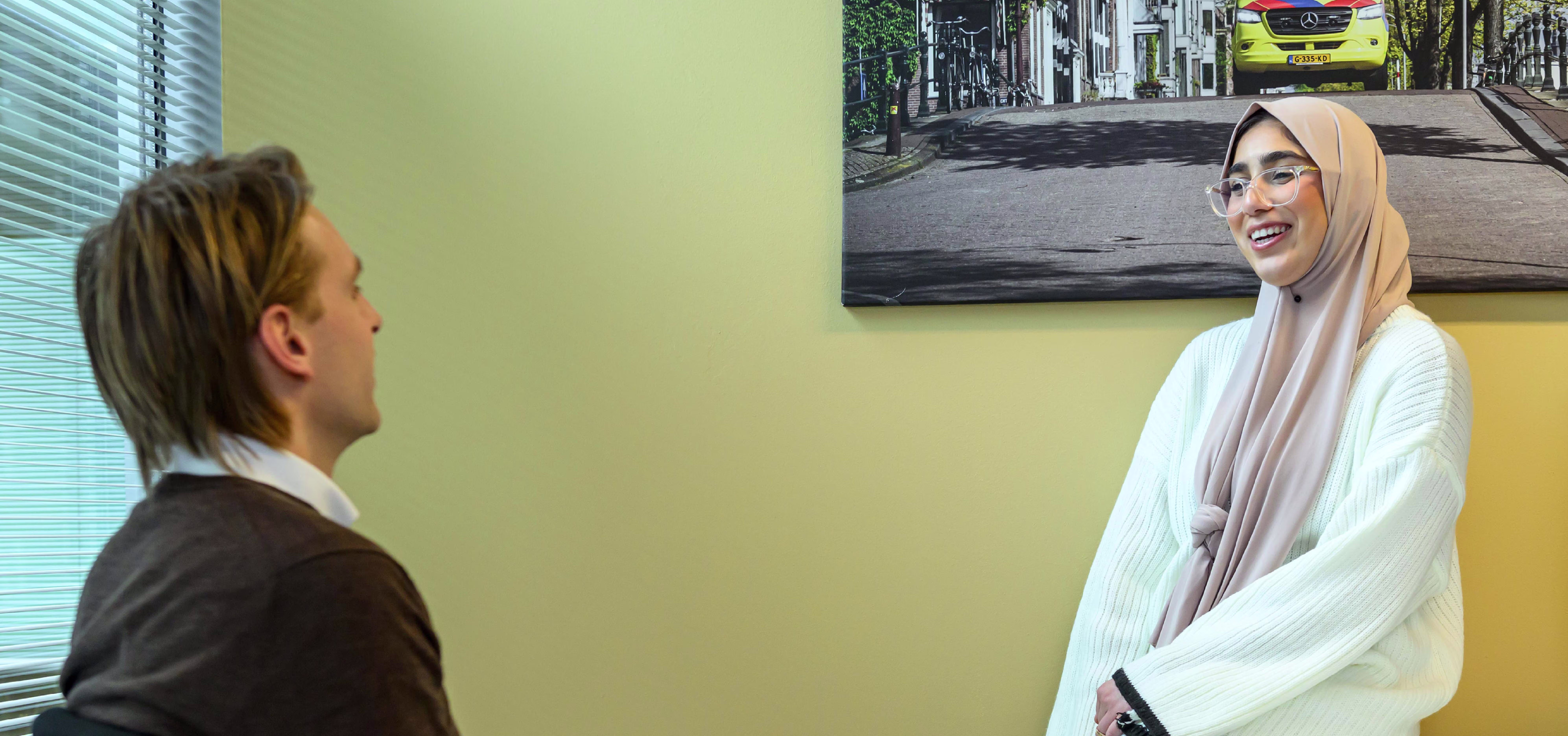 Twee collega's in gesprek op kantoor, een man in pak en een vrouw met hoofddoek in witte trui, bij raam met jaloezie.