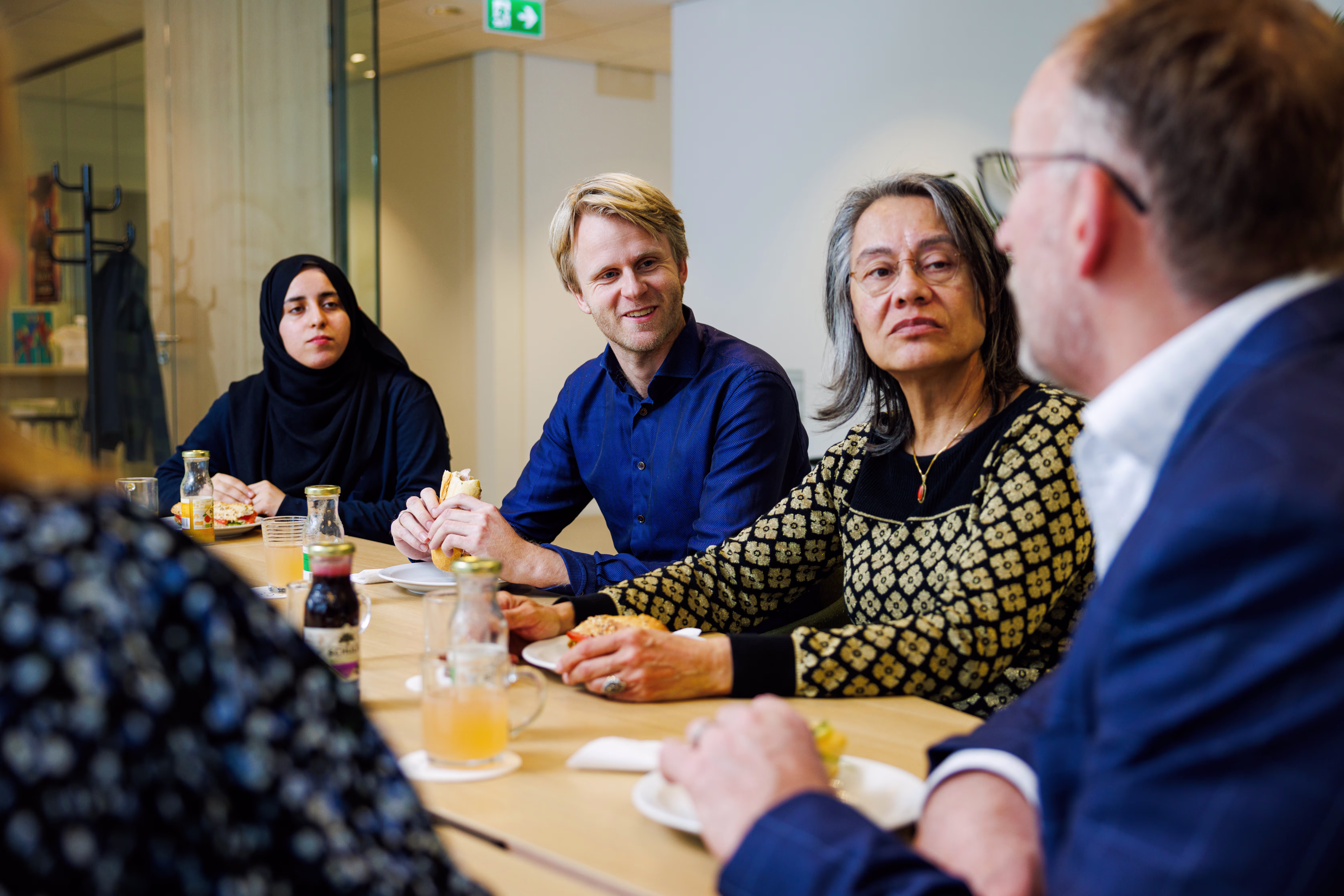 Diverse groep collega's zit rond tafel tijdens zakelijke lunch of vergadering in modern kantoor.