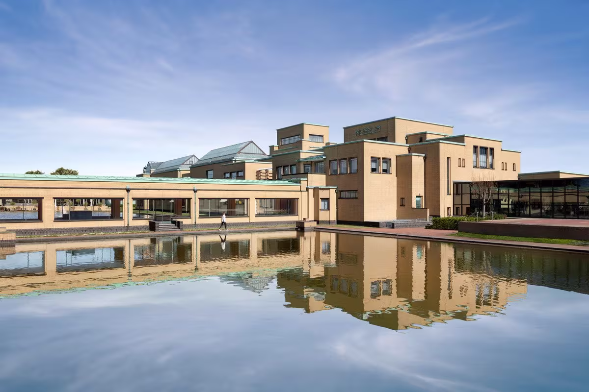 Modern museumgebouw met beige gevel weerSpiegeld in water, onder blauwe hemel met witte wolken.