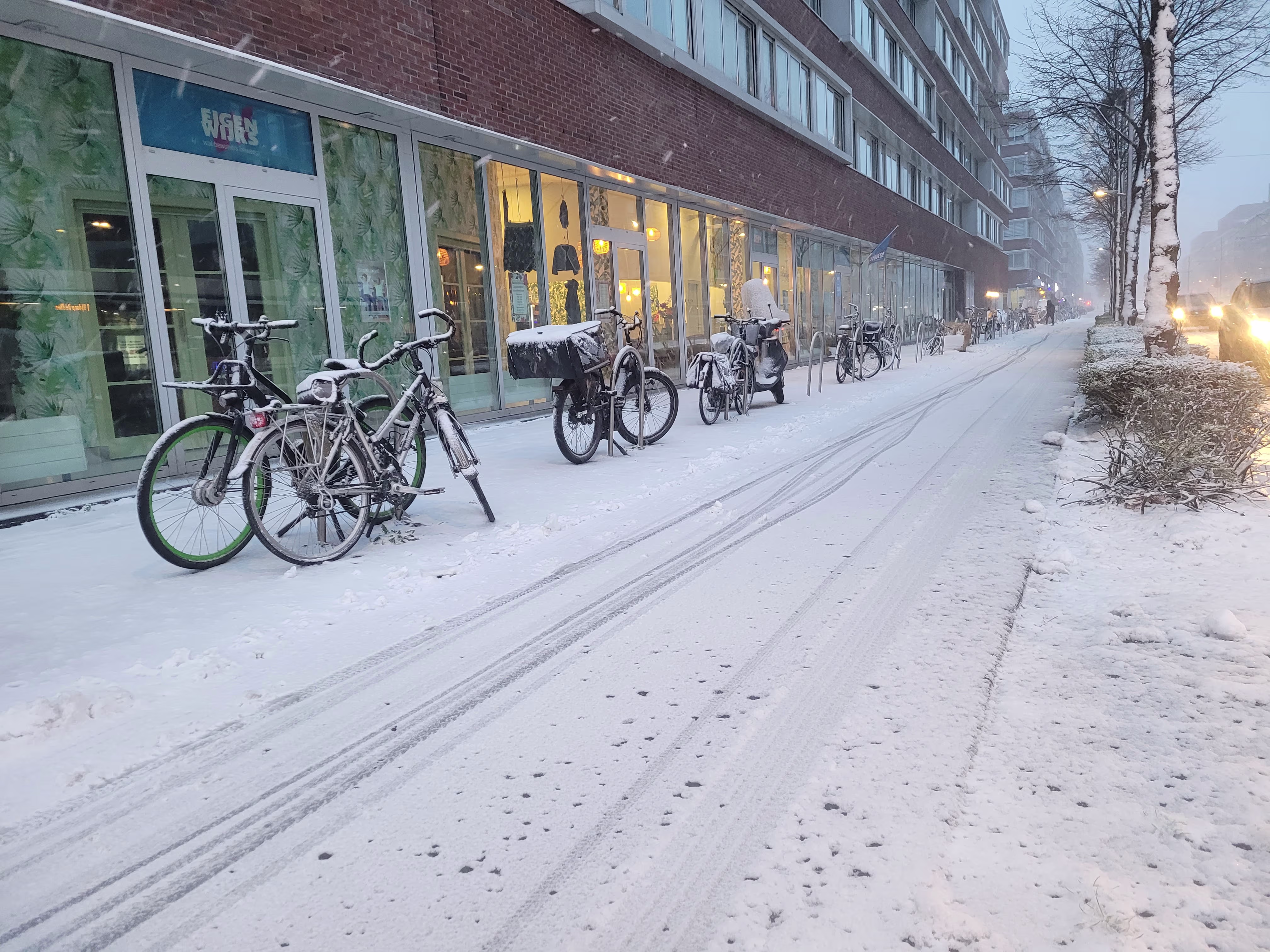 Besneeuwde straat met fietsen voor winkels en appartementen op een winterse avond.