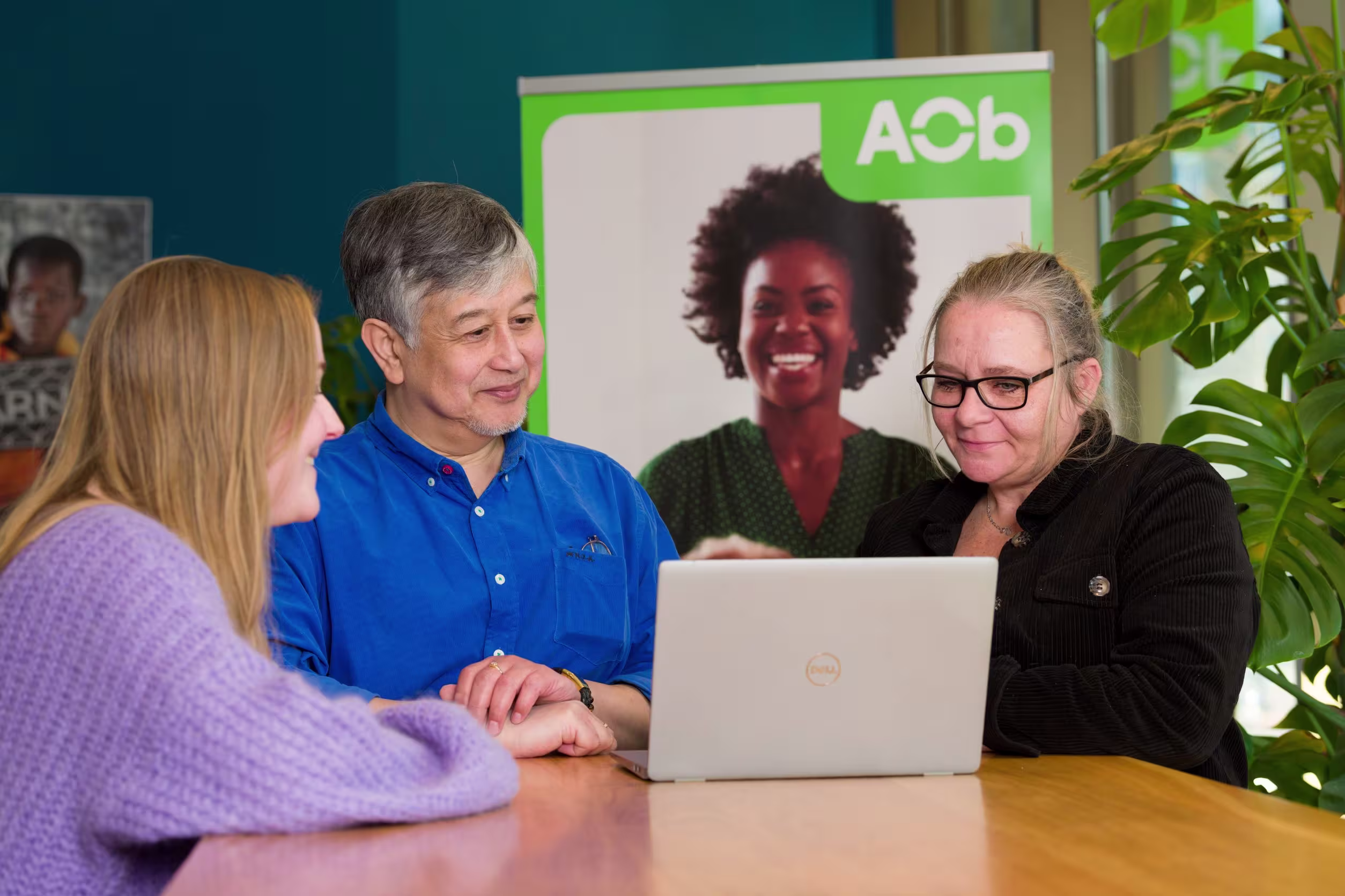 Drie collega's bespreken samen iets op een laptop aan een houten tafel, met een groen AOb-bord op de achtergrond.