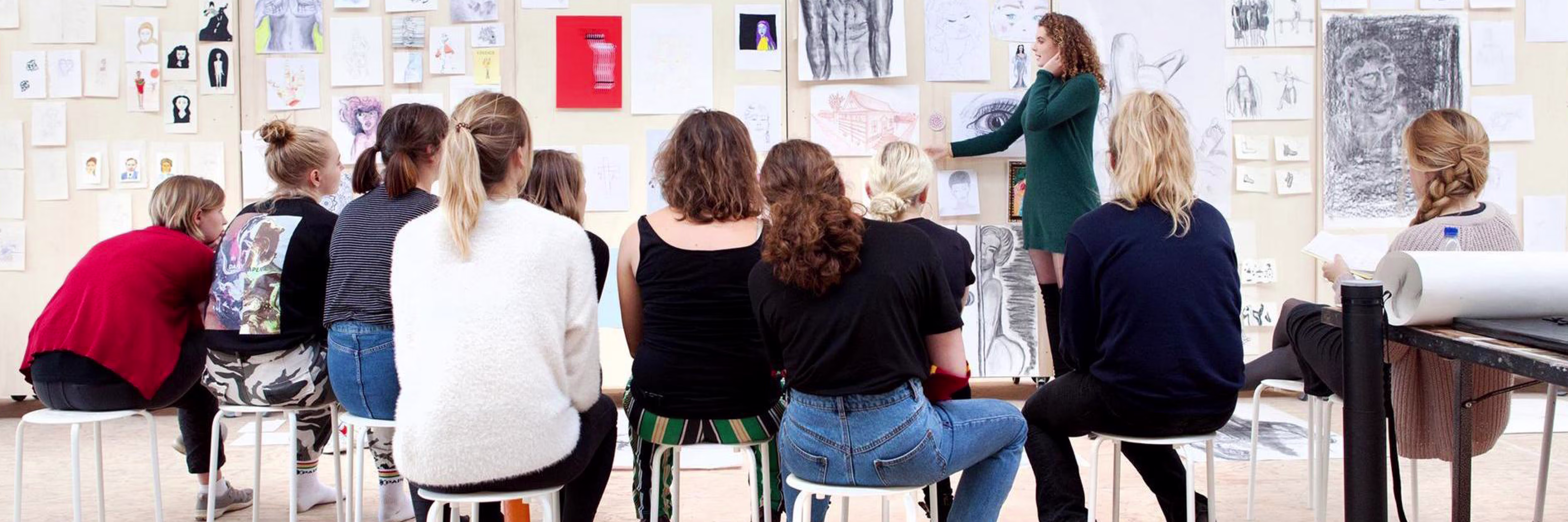 Een docent geeft les aan een groep studenten die op krukken zitten voor een muur vol kunstwerken en tekeningen.
