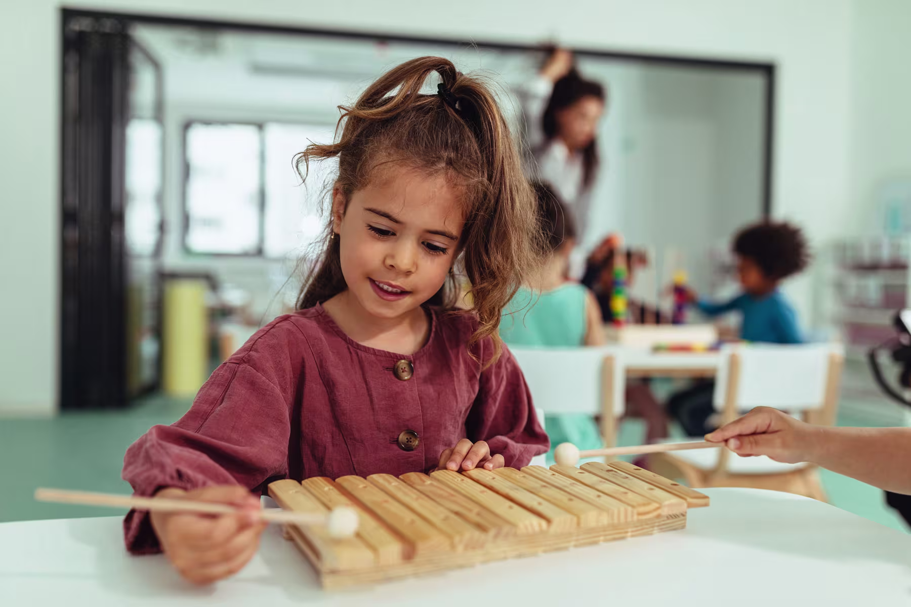 Een meisje speelt met een houten xylofoon in een moderne klaslokaal, terwijl andere kinderen op de achtergrond bezig zijn.