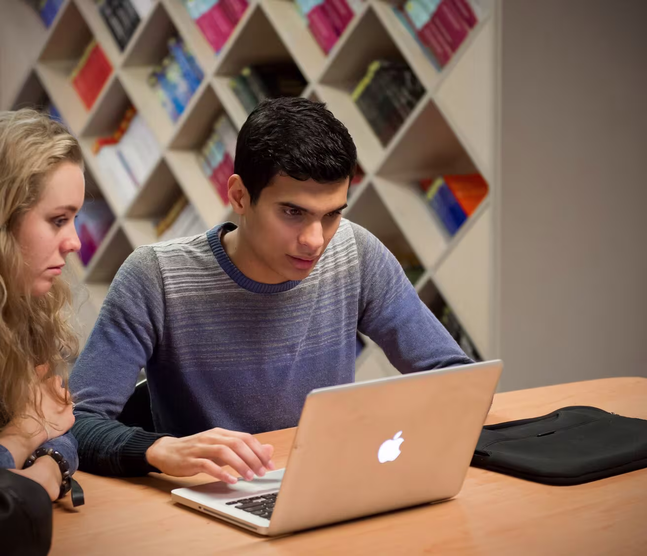 Twee studenten werken samen achter een laptop in een bibliotheek met boekenkasten op de achtergrond.
