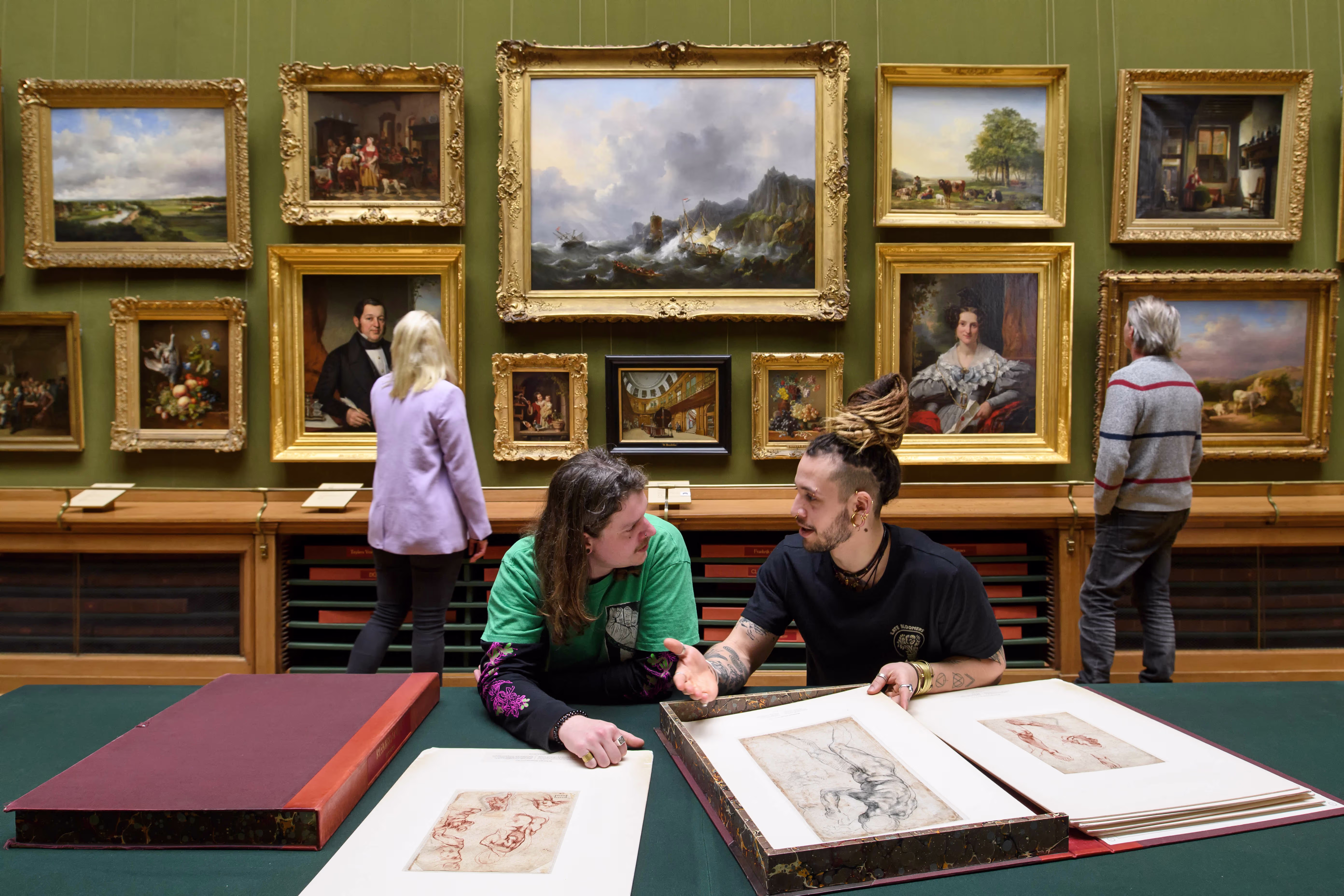 Twee mensen bekijken tekeningen in een museum, met klassieke schilderijen aan de groene muur op de achtergrond.