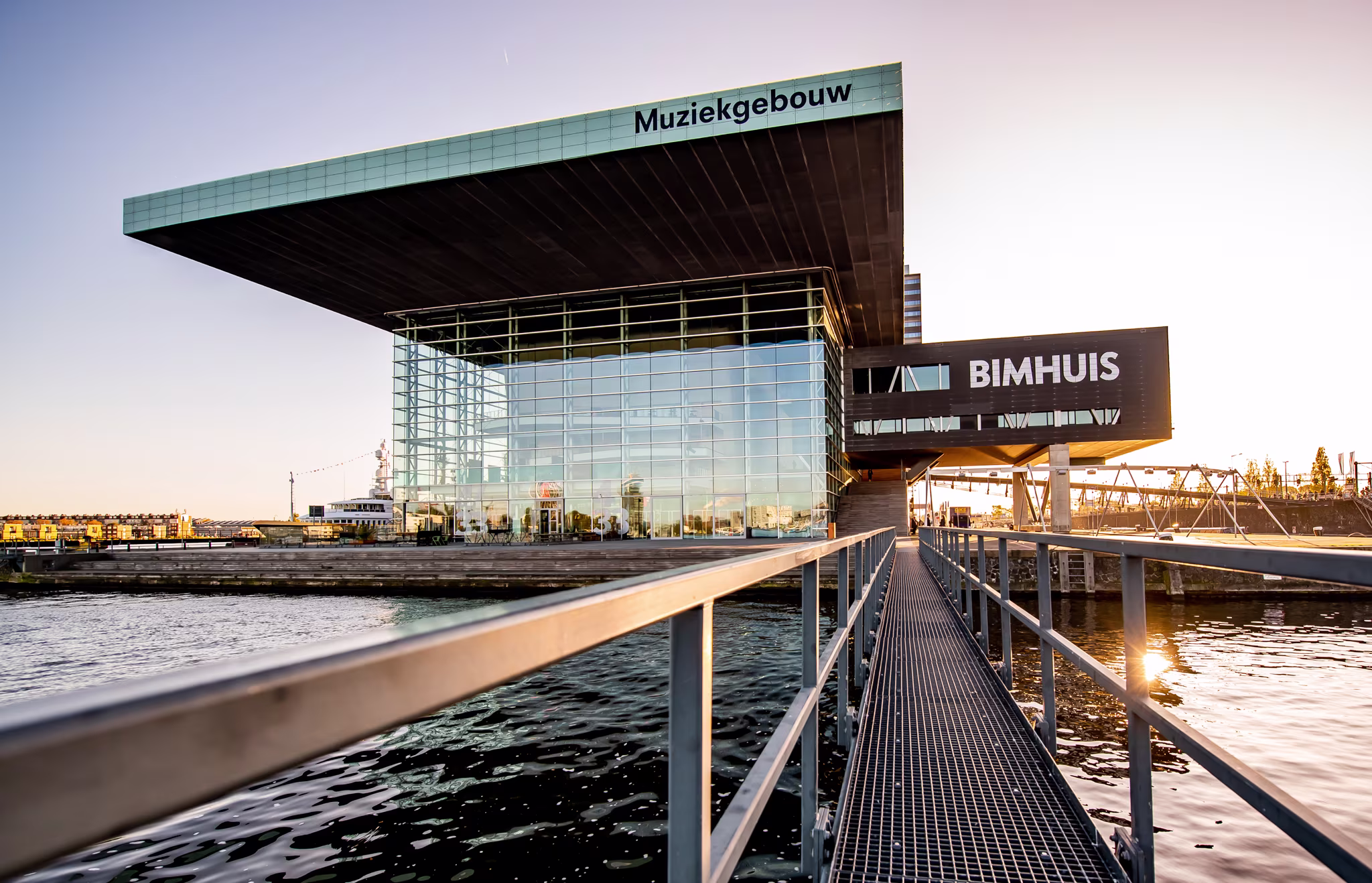 Modern glazen gebouw van Muziekgebouw aan 't IJ met aangrenzend Bimhuis, gezien vanaf een loopbrug over het water bij zonsondergang.