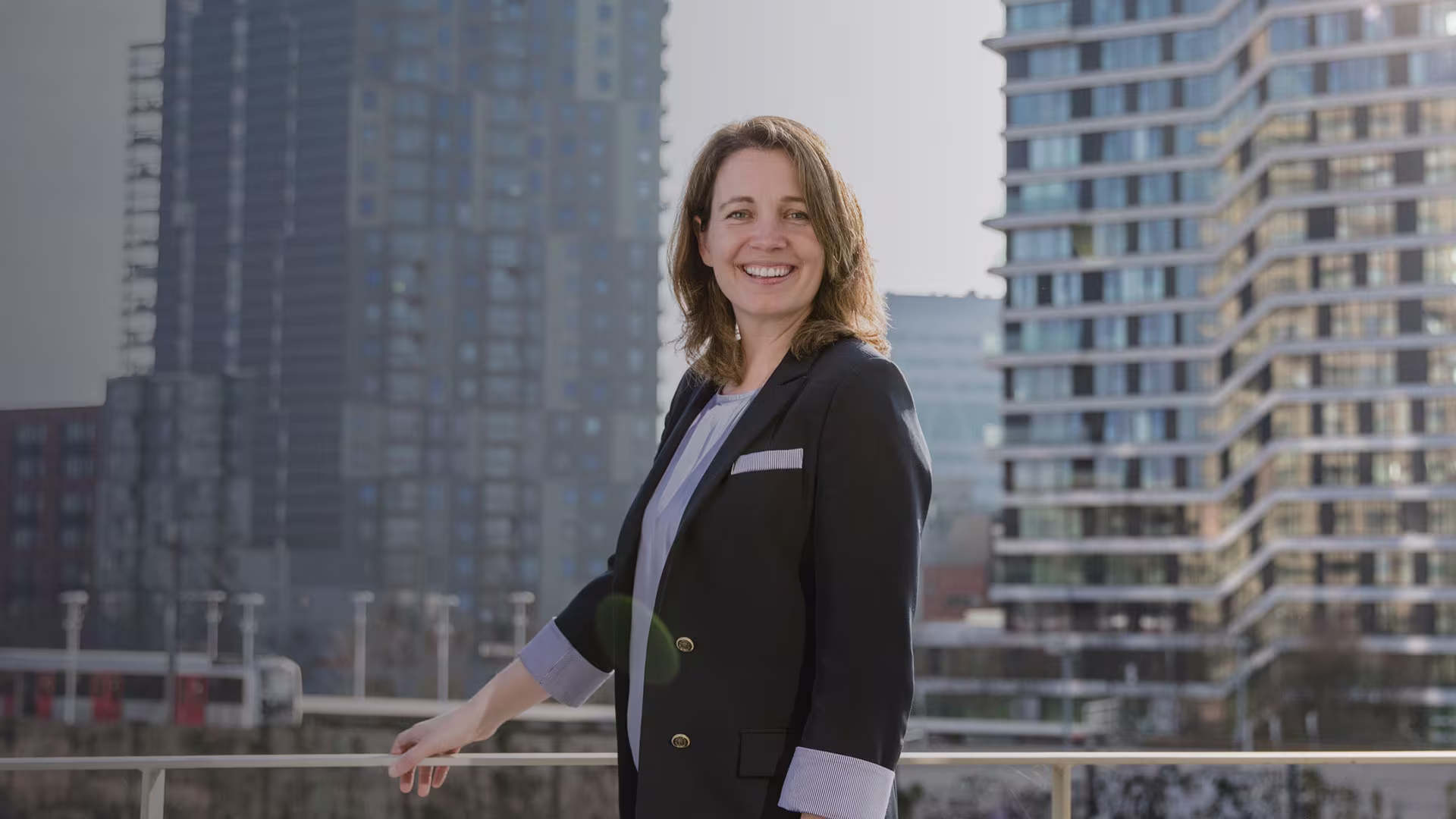 Vrouw in donker zakelijk jasje lacht vriendelijk op kantoorbalkon met moderne hoogbouw op de achtergrond.