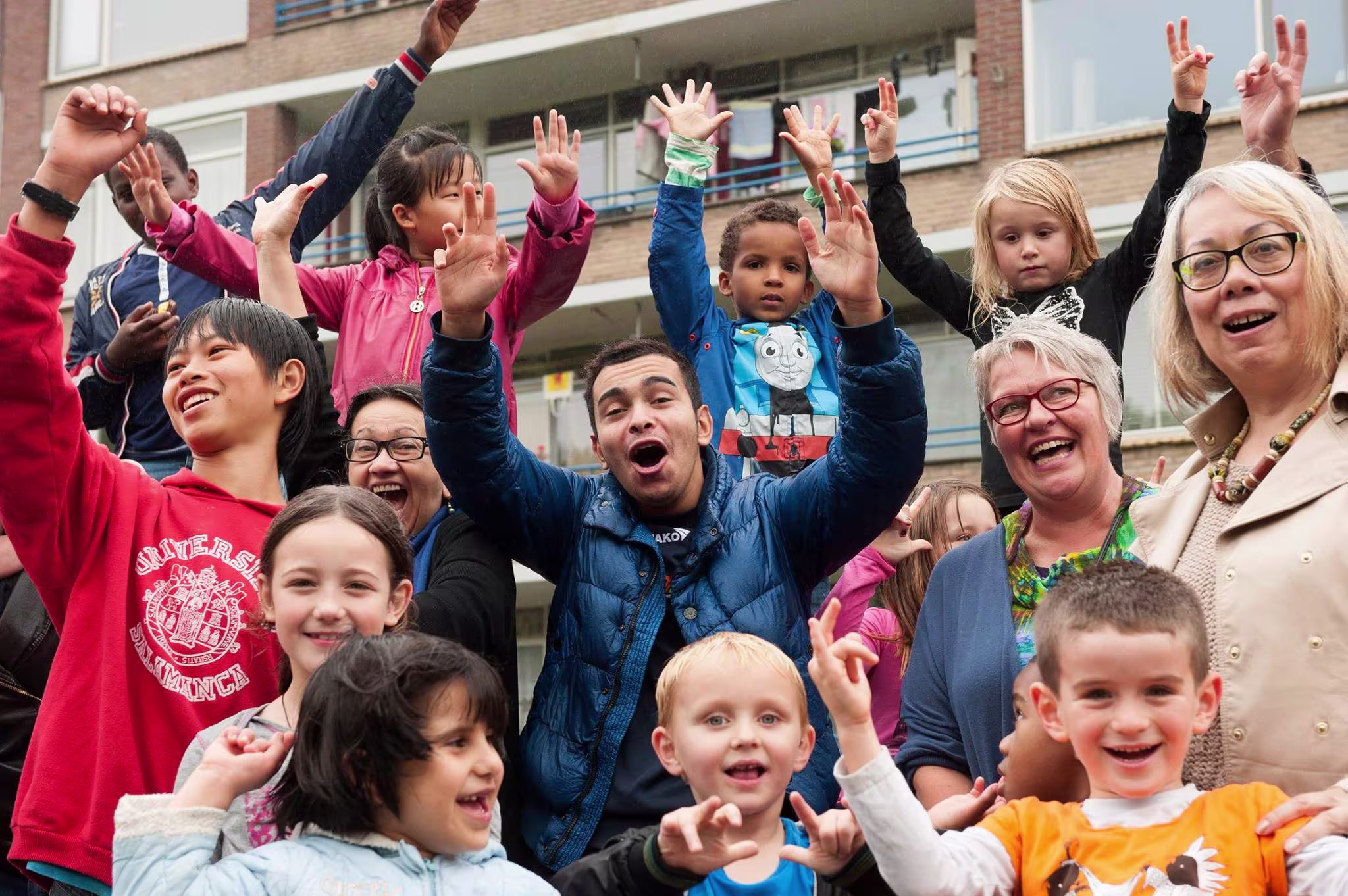 Een groep kinderen en begeleiders juichen enthousiast met hun handen omhoog buiten voor een gebouw.