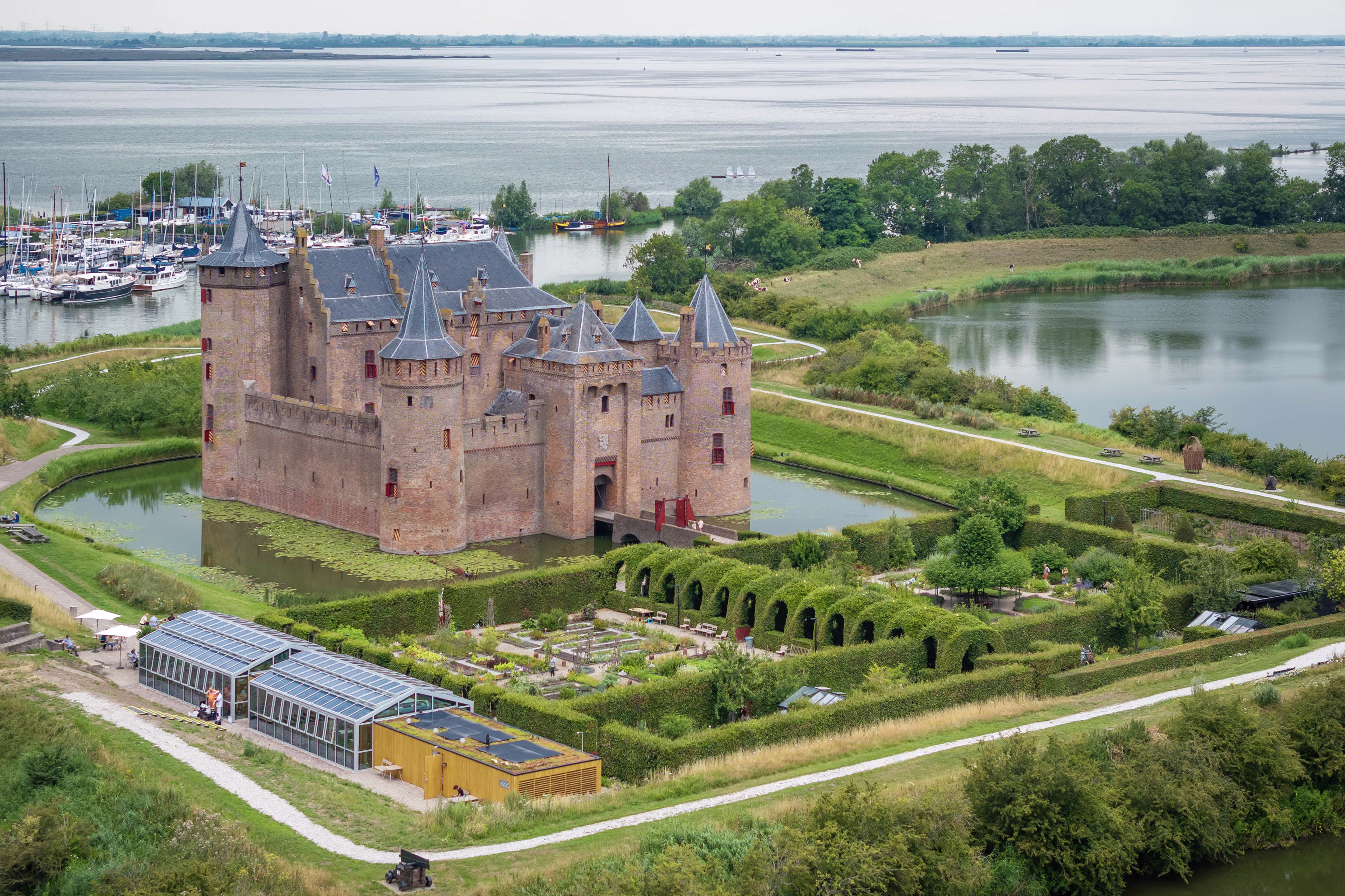 Luchtfoto van het middeleeuwse Muiderslot omringd door water en groene tuinen, met een jachthaven op de achtergrond.
