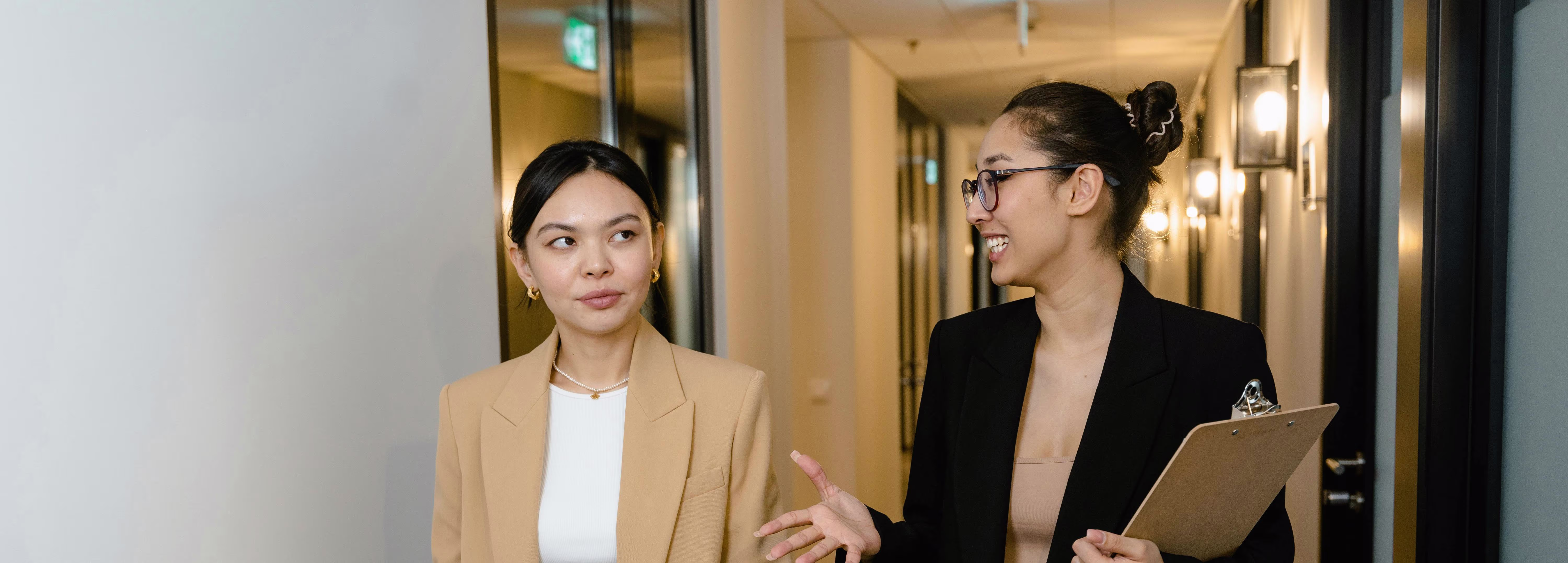Twee professioneel geklede vrouwen lopen pratend door een moderne gang, een draagt een clipboard.