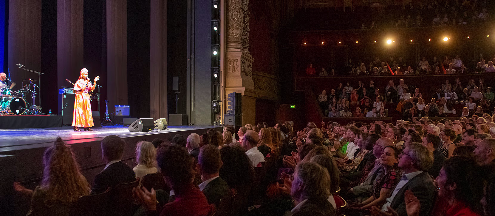Een artiest in een witte jurk staat op het podium van een theater vol publiek tijdens een Holland Festival voorstelling.