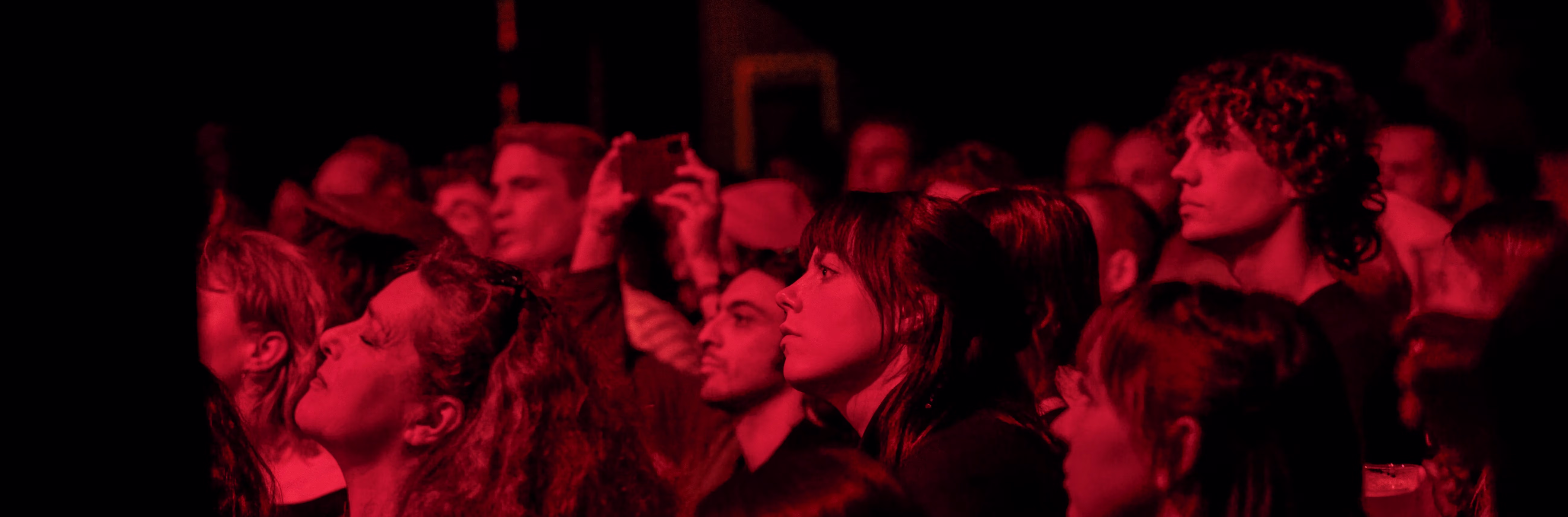 Een publiek kijkt geconcentreerd toe tijdens een voorstelling, verlicht door warm rood licht in een donkere ruimte.