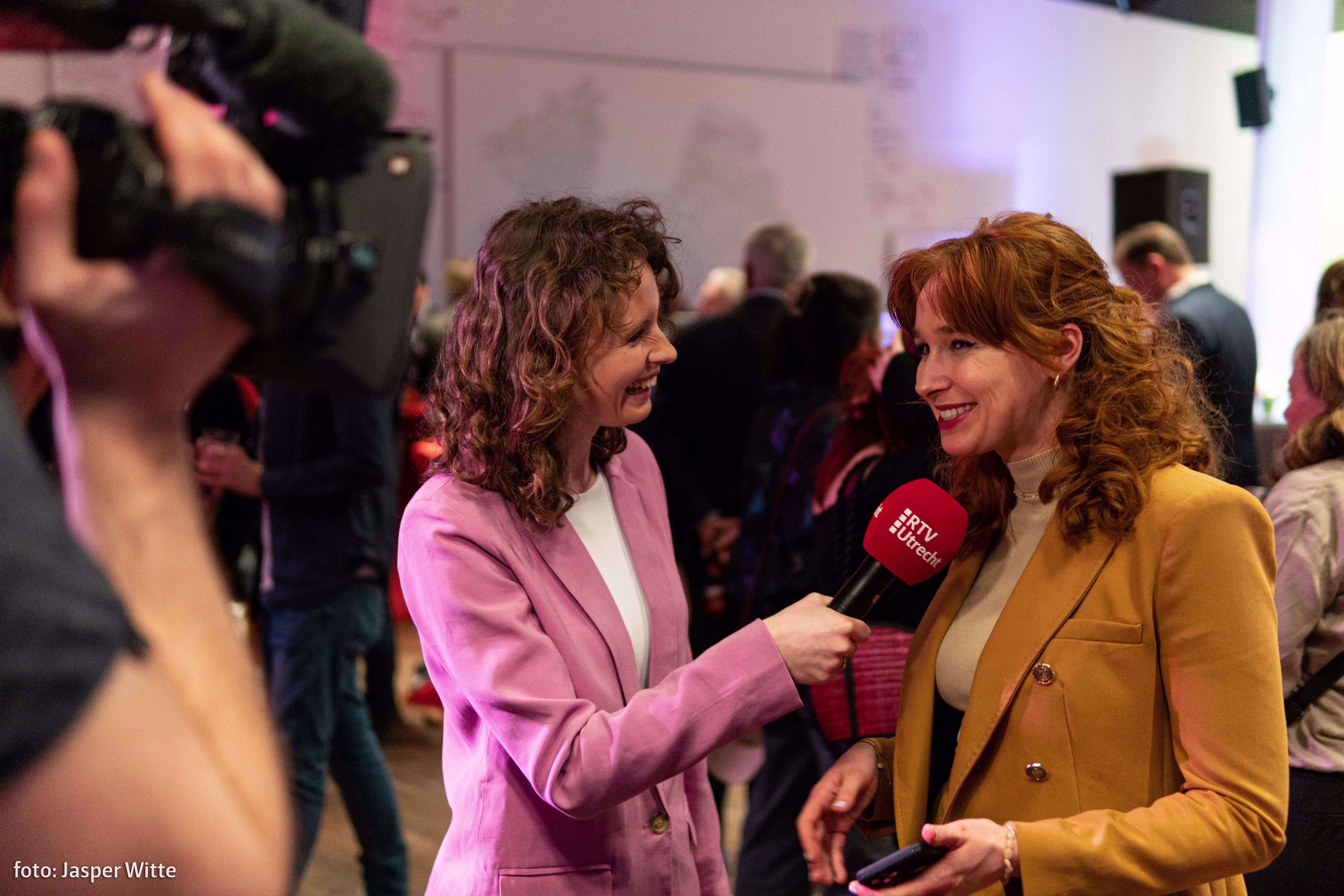 Een journaliste in roze pak interviewt een vrouw in gele jas met een rode microfoon tijdens een evenement met camera's op de achtergrond.