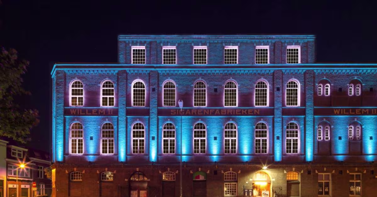 Een historisch bakstenen gebouw met blauwe verlichting 's avonds, met de tekst "Willem Twee" op de gevel.