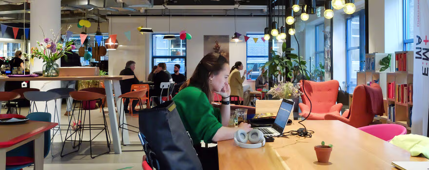 Een moderne kantoorruimte met medewerkers die werken aan laptops aan kleurrijke tafels, met industrieel plafond en grote ramen.