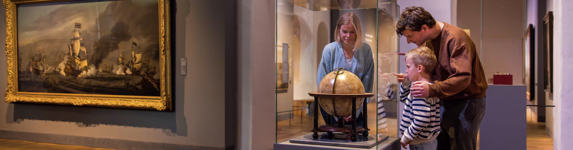 Vader en kind bekijken een globe in het Scheepvaartmuseum, met zeilschip schilderij op de achtergrond.