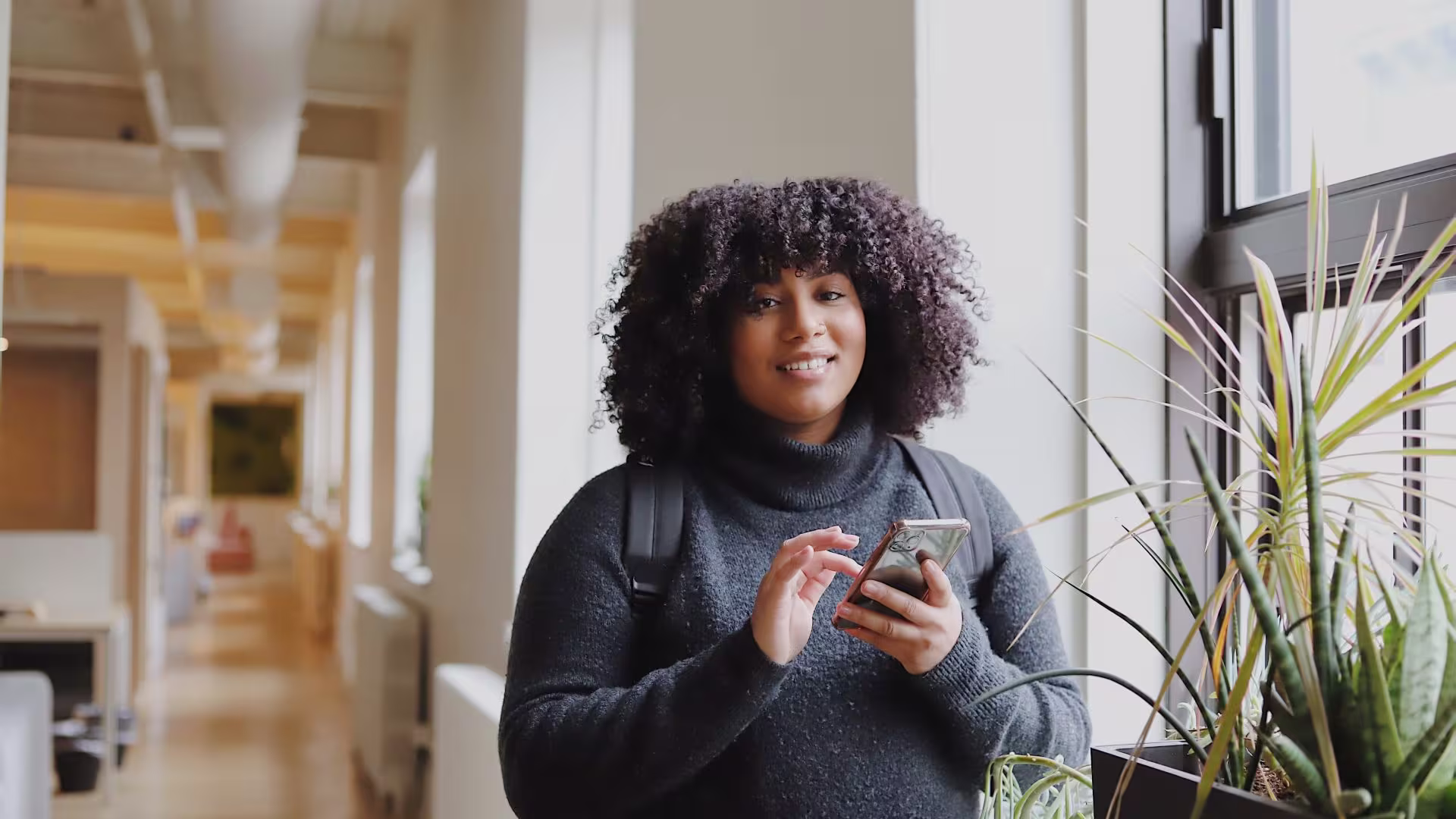 Een glimlachende vrouw met krullend haar houdt een telefoon vast in een modern kantoor met planten op de achtergrond.