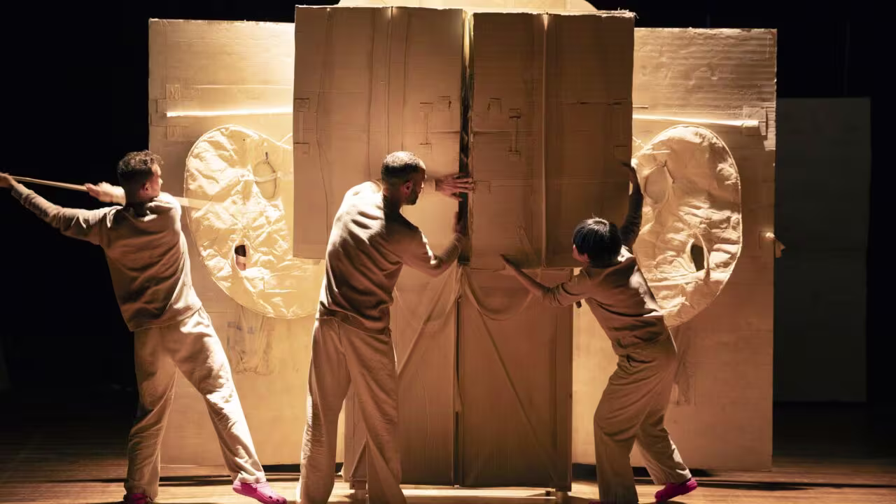 Drie acteurs in bruine gewaden duwen tegen grote kartonnen decorstukken op een theaterpodium tijdens een voorstelling.