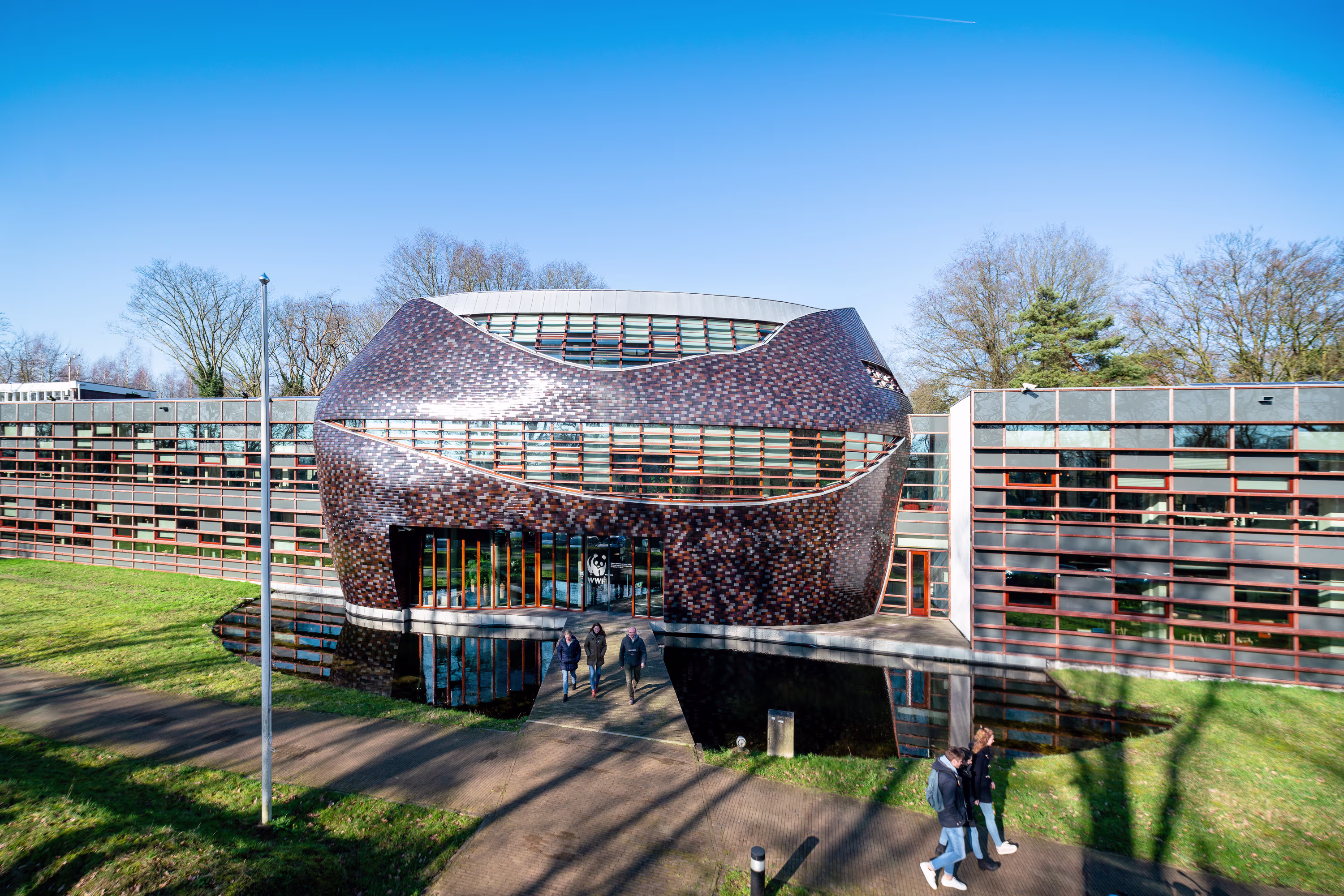 Modern gebouw met organische vorm en kleurrijke gevel, omgeven door groen en wandelpaden met bezoekers.