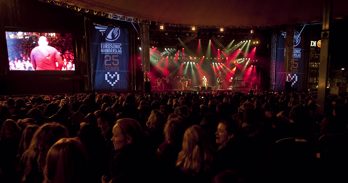 Een grote concertzaal vol publiek kijkt naar een podium met artiesten, verlicht door kleurrijke spotlights en grote schermen.