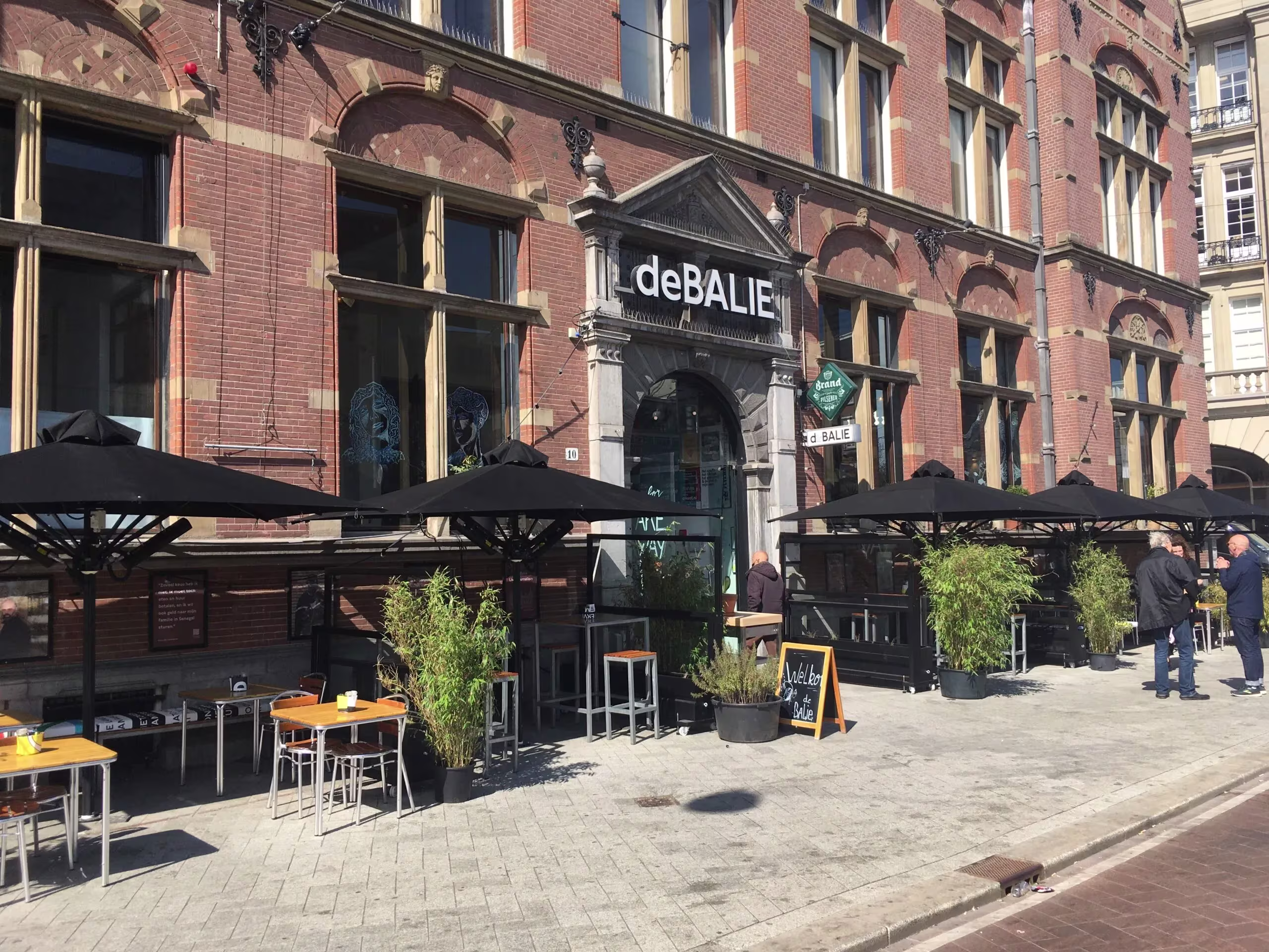 Buitenkant van theater De Balie in Amsterdam met rood bakstenen gevel en terras met zwarte parasols.