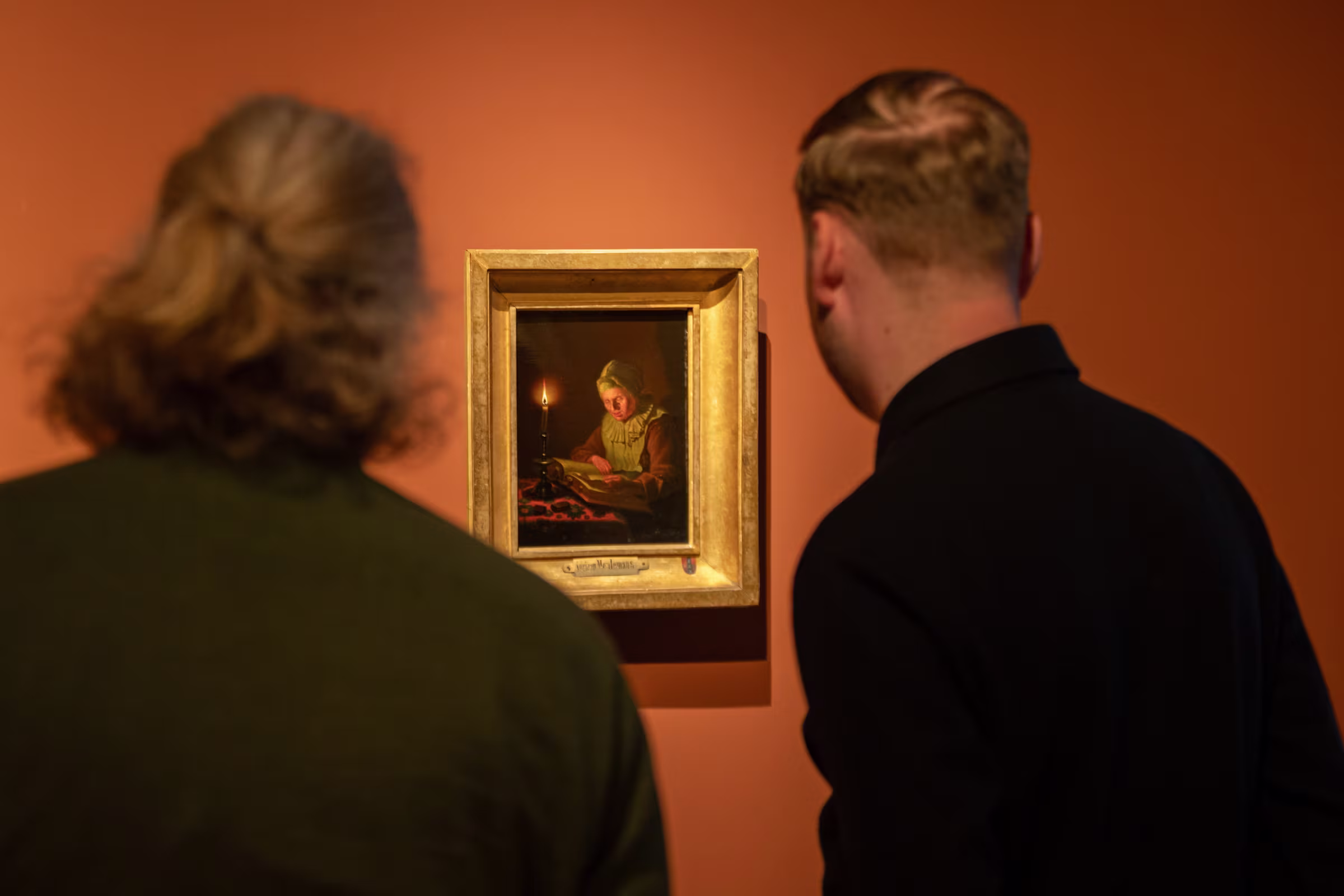 Twee bezoekers bekijken een schilderij in gouden lijst met kaarslicht tafereel aan oranje museumwand.