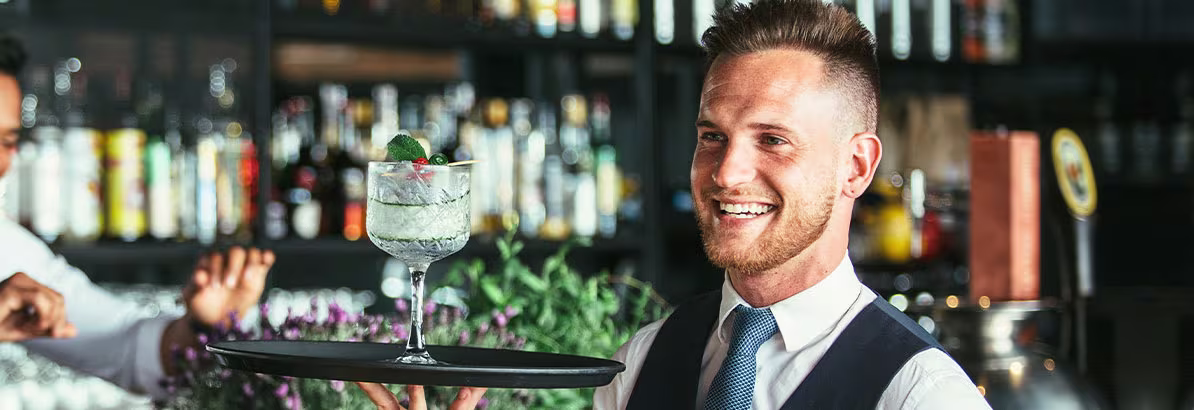 Een glimlachende ober in vest en stropdas serveert een cocktail in een bar met flessen op de achtergrond.