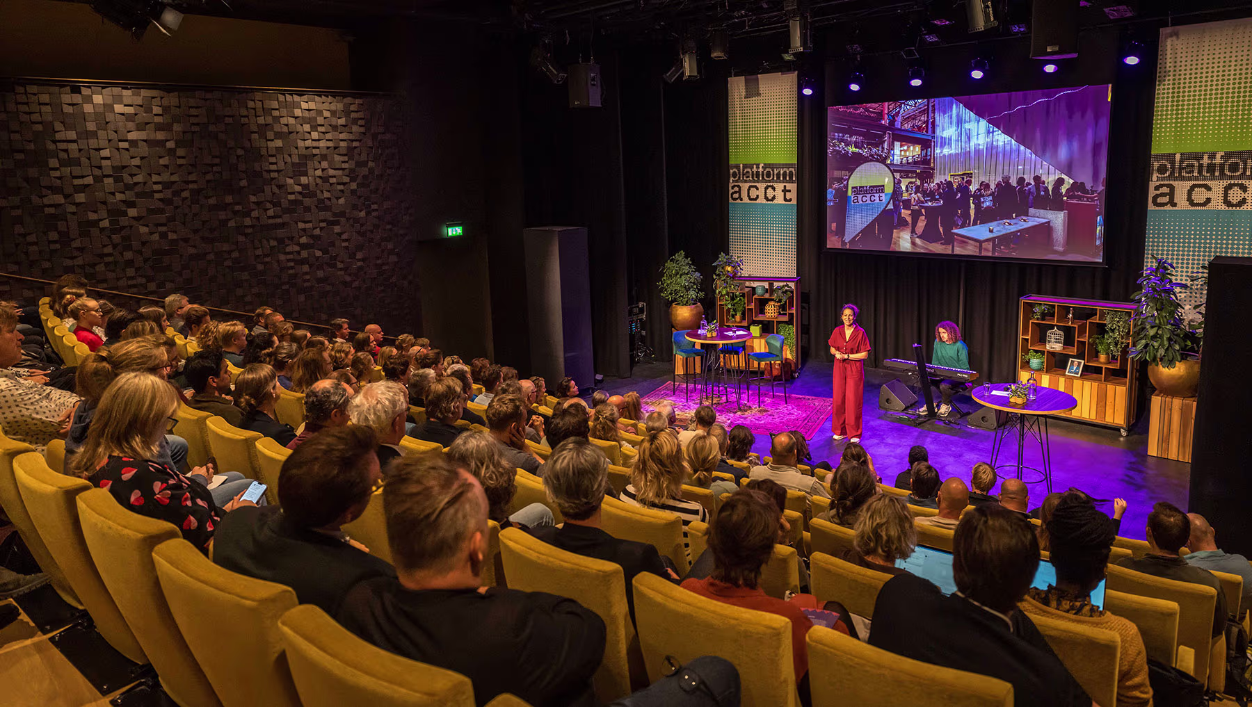 Een volle theaterzaal met gele stoelen waar publiek luistert naar een presentatie op het podium met paarse verlichting.